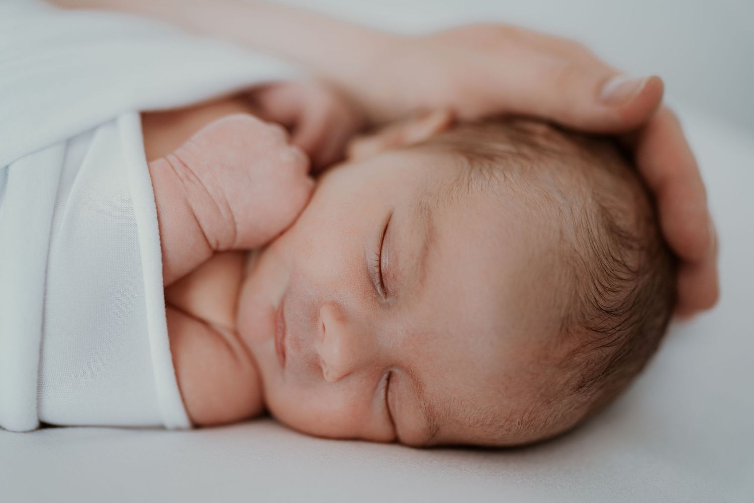 A peaceful newborn baby sleeps soundly while swaddled in a soft white blanket against a light background.