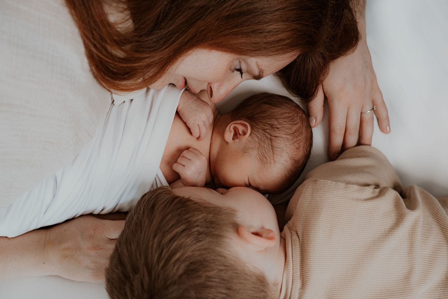 A close-up intimate photo shows three people snuggling together on white bedding in soft natural lighting.