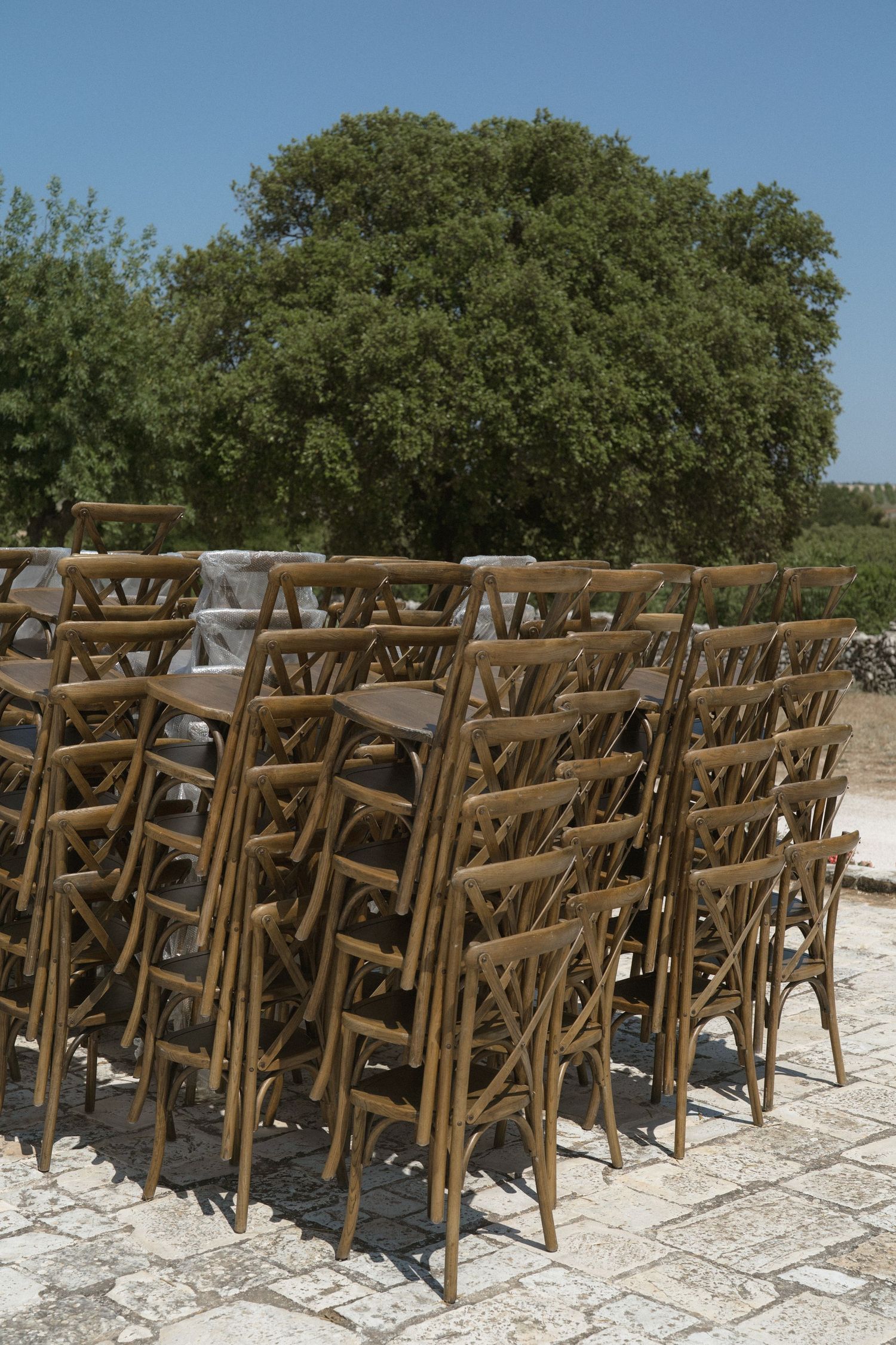 Multiple rows of rustic wooden chairs set up for an outdoor event on a stone patio surrounded by green trees.