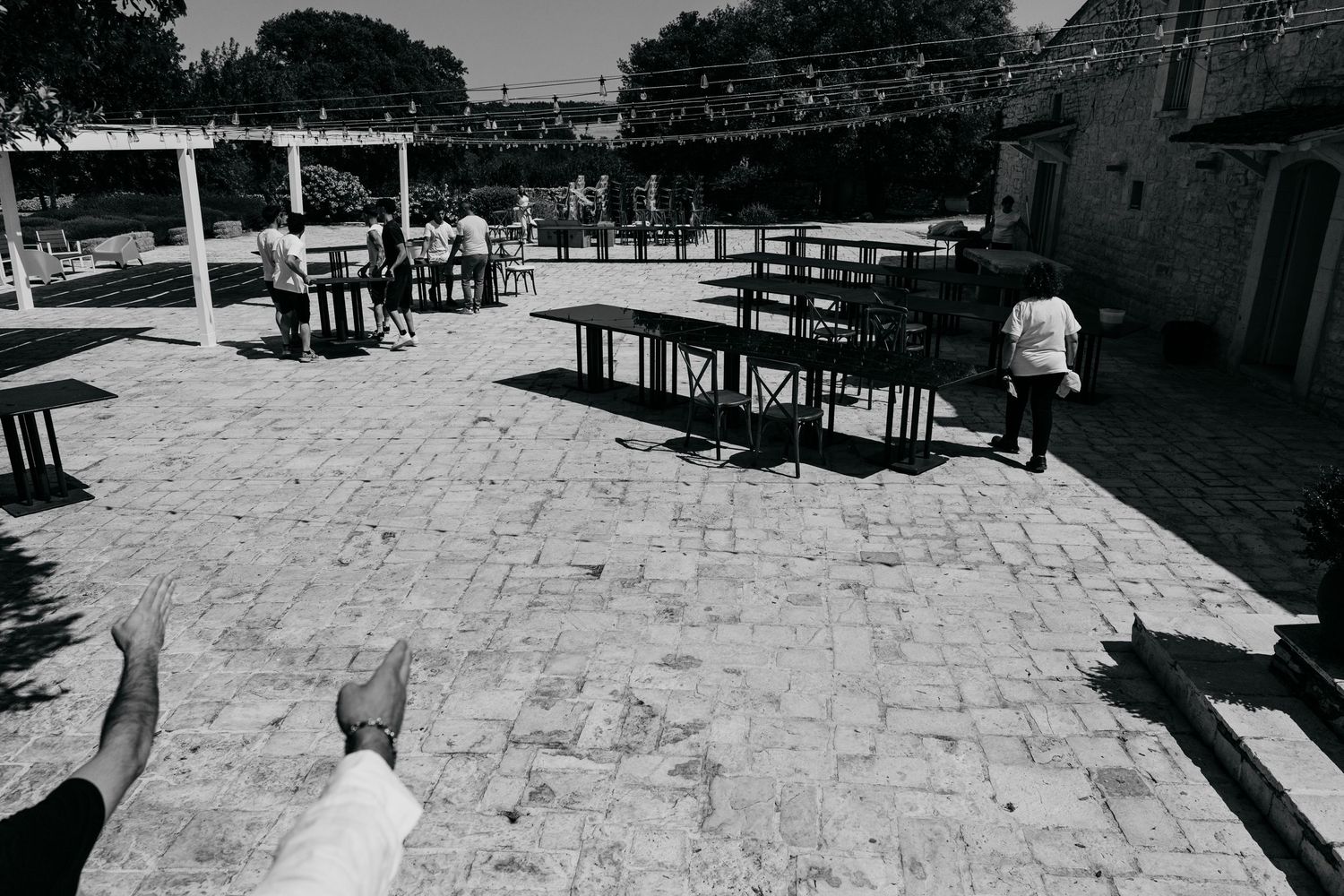 A group of people relax at outdoor dining tables on a sunny patio with stone tiled flooring.