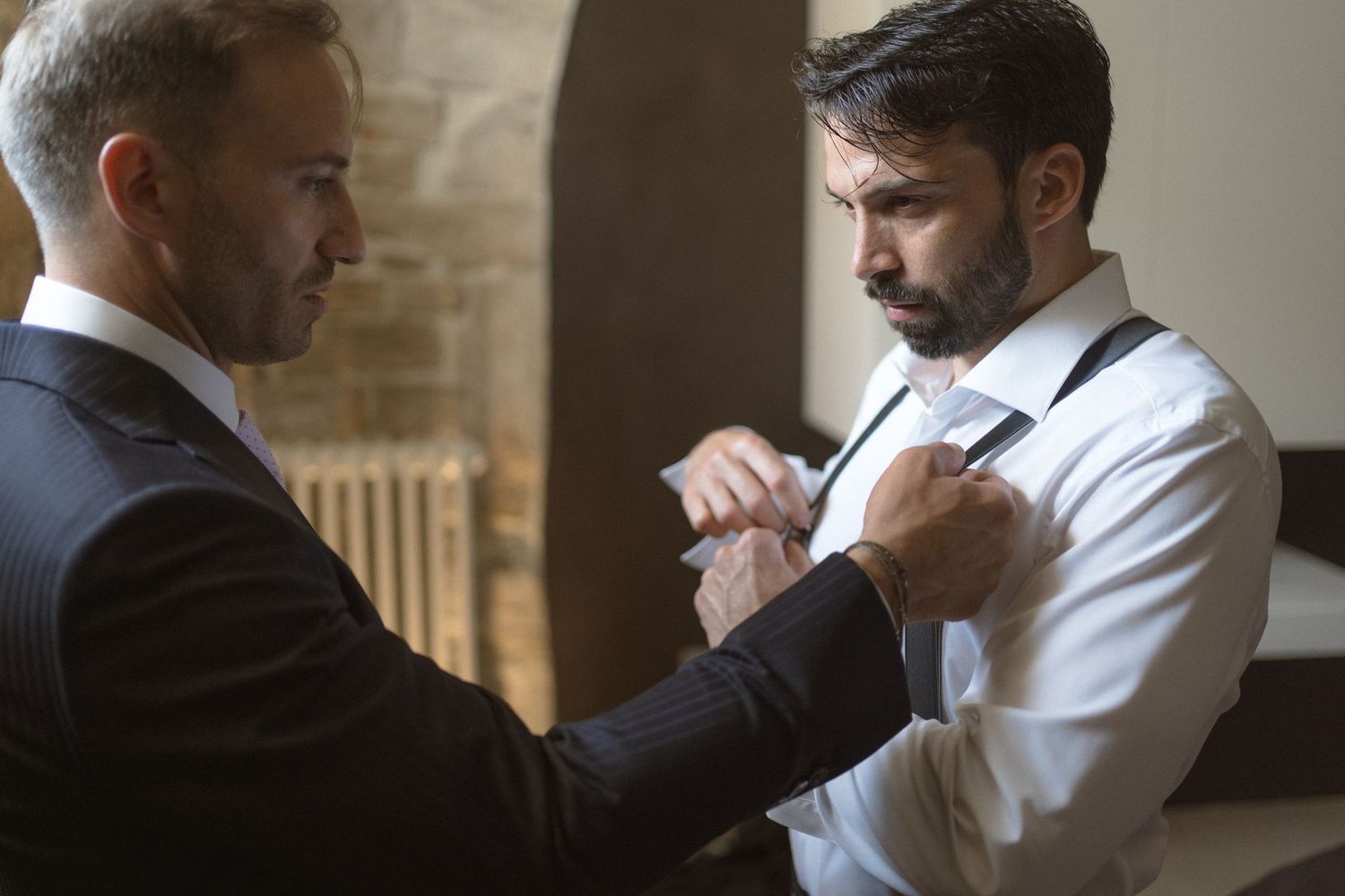 Groomsmen help adjust formal attire before wedding ceremony in stone villa interior.
