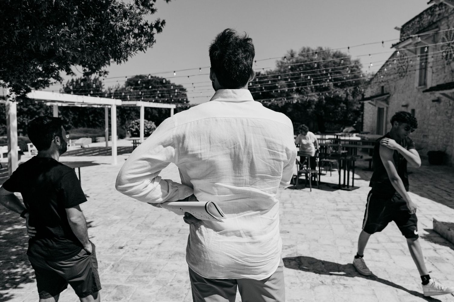 A black and white photograph shows people gathered in a sunny outdoor courtyard space during daytime.