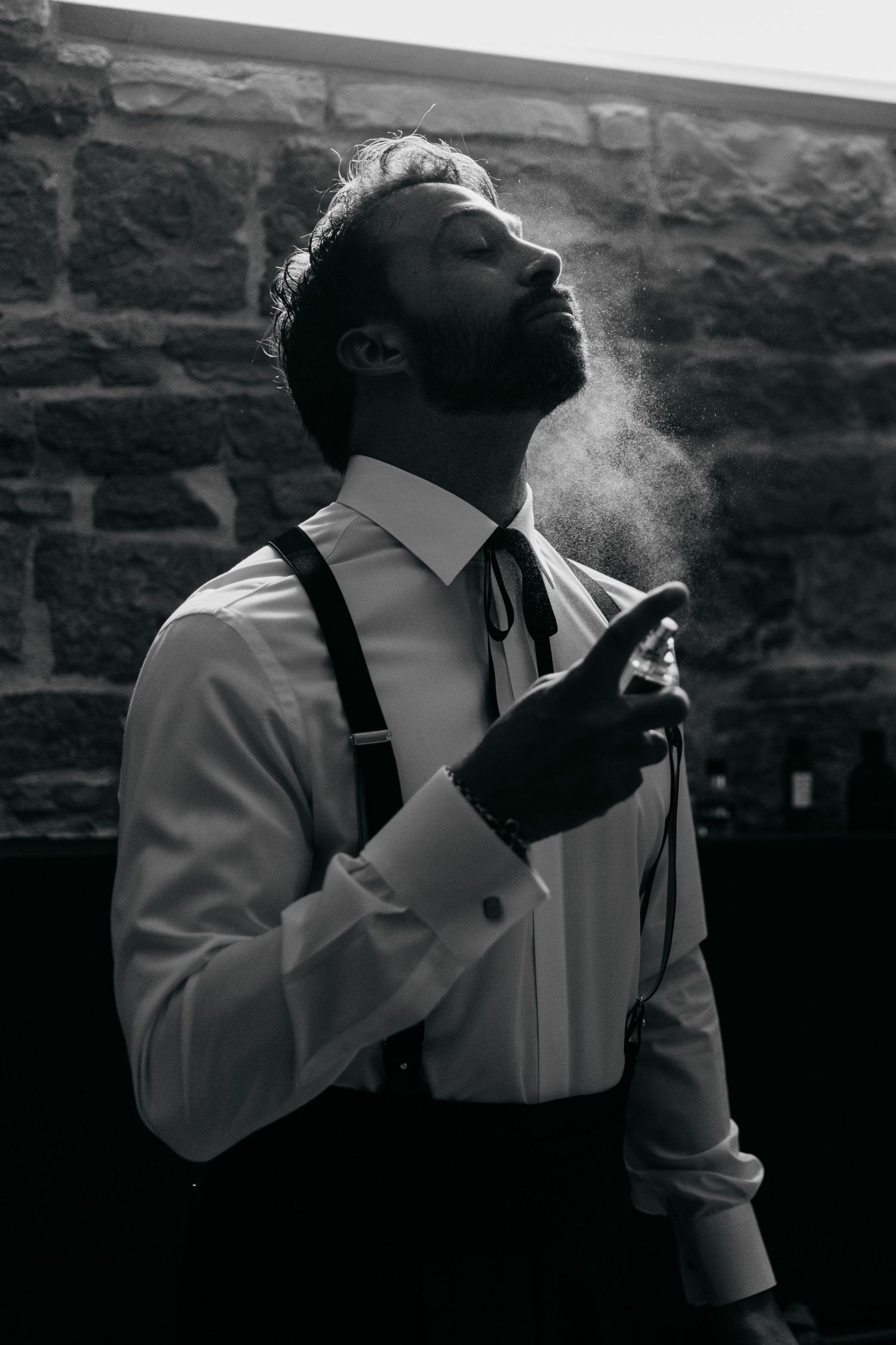 Dramatic black and white photos of a man in suspenders smoking against a brick wall.