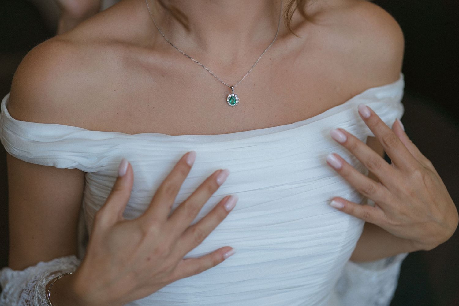 Close up detail of white off-shoulder wedding dress with delicate necklace and manicured hands.