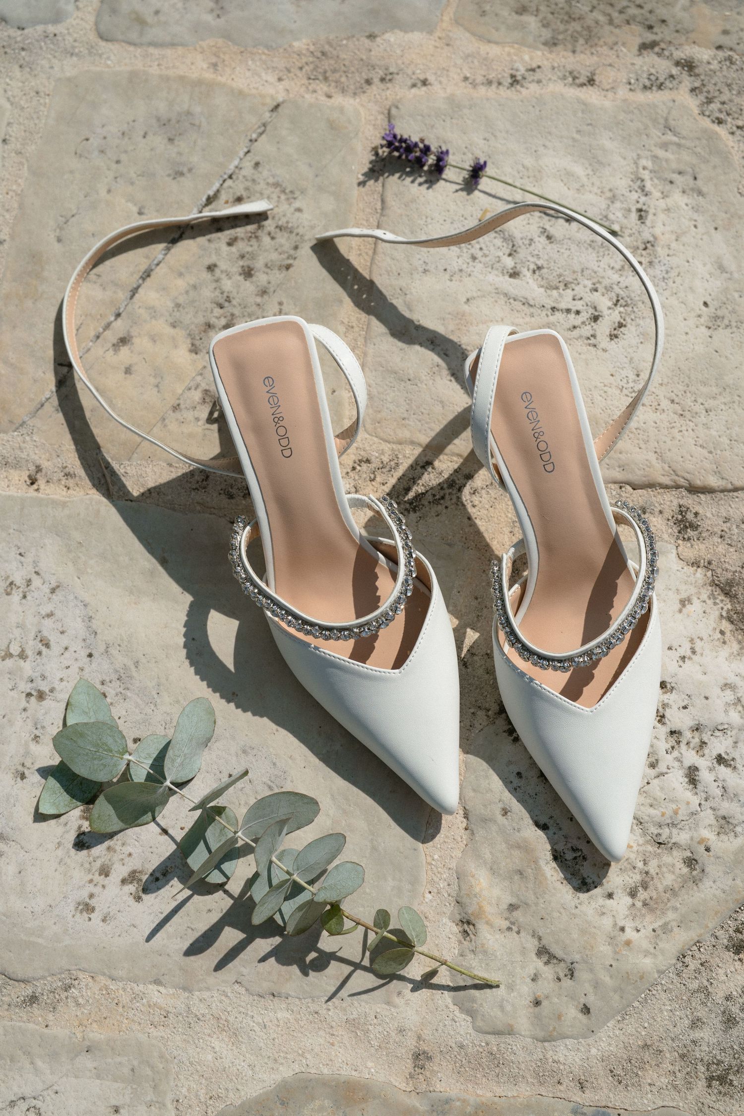 Elegant white bridal shoes with eucalyptus leaves on a stone surface in natural light.