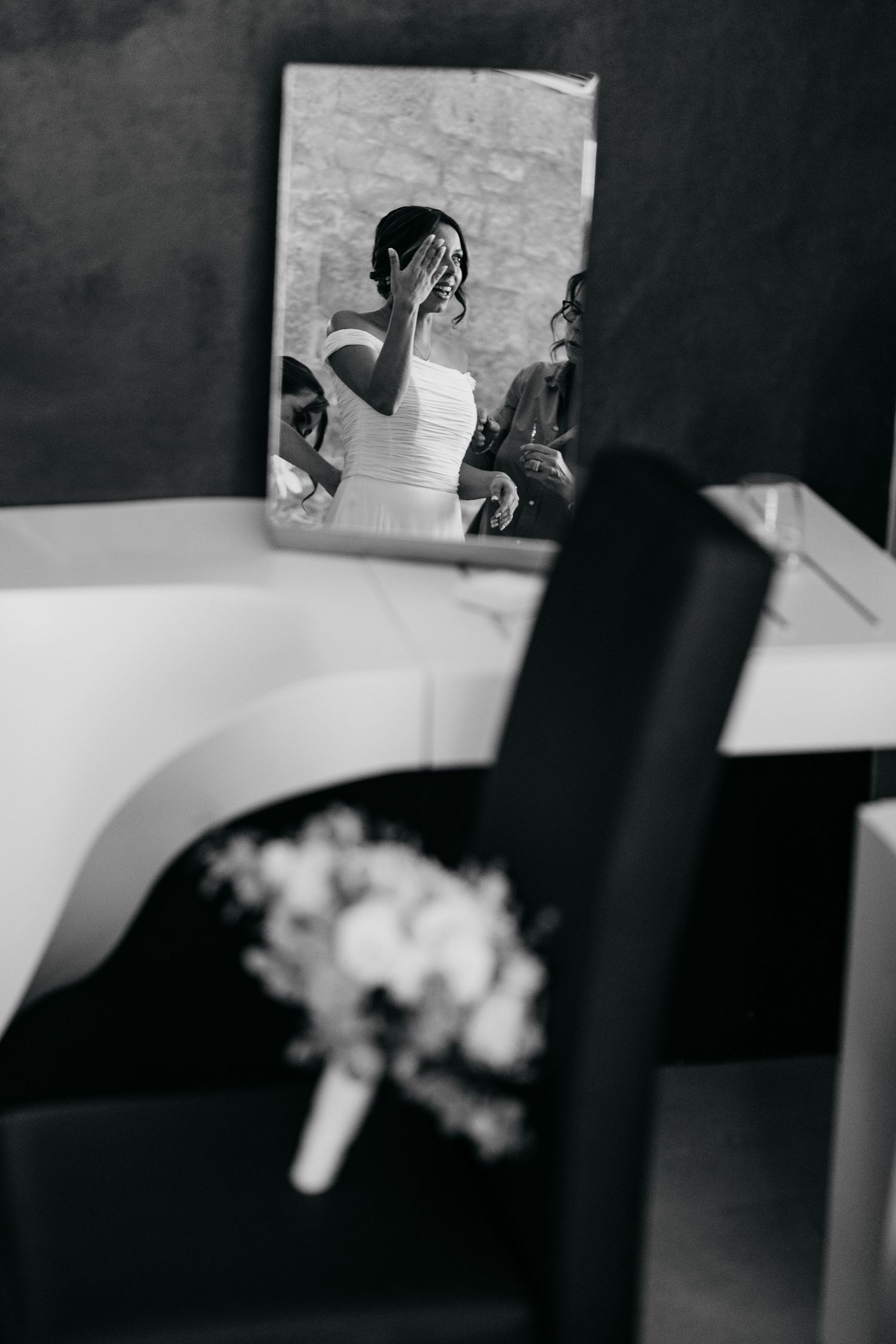 Artistic black and white reflection of a bride in a wide mirror with a bouquet in the foreground.