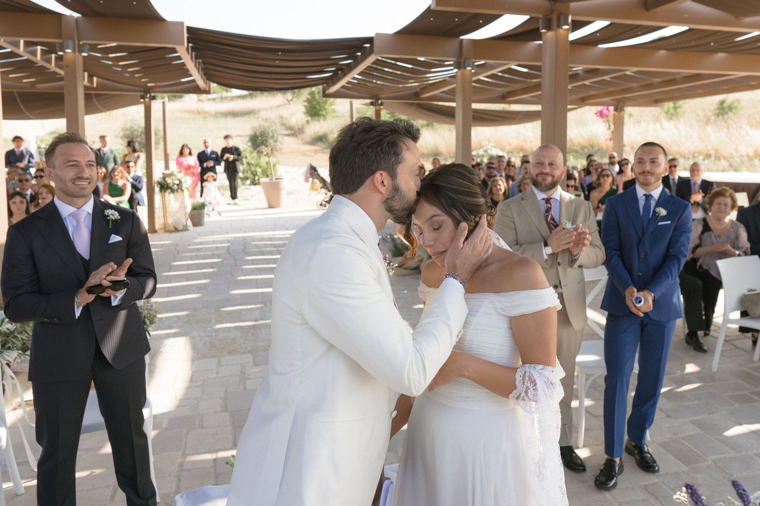 Outdoor wedding celebration under wooden pergola with string lights and guests mingling.