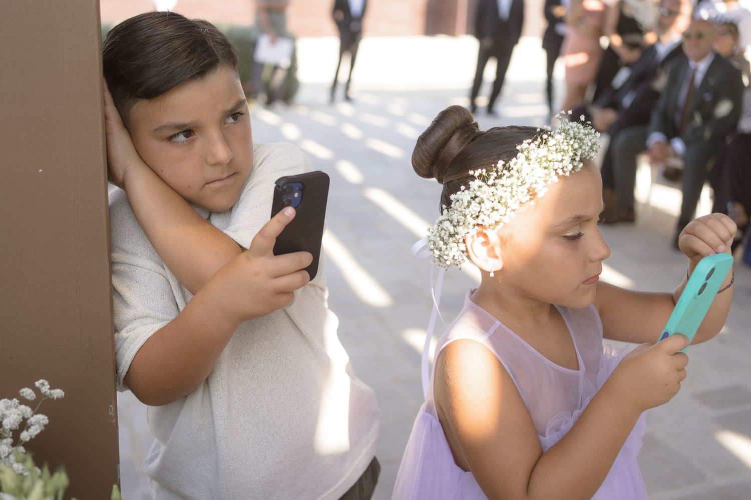 Two children using mobile phones while dressed in formal attire at an event.