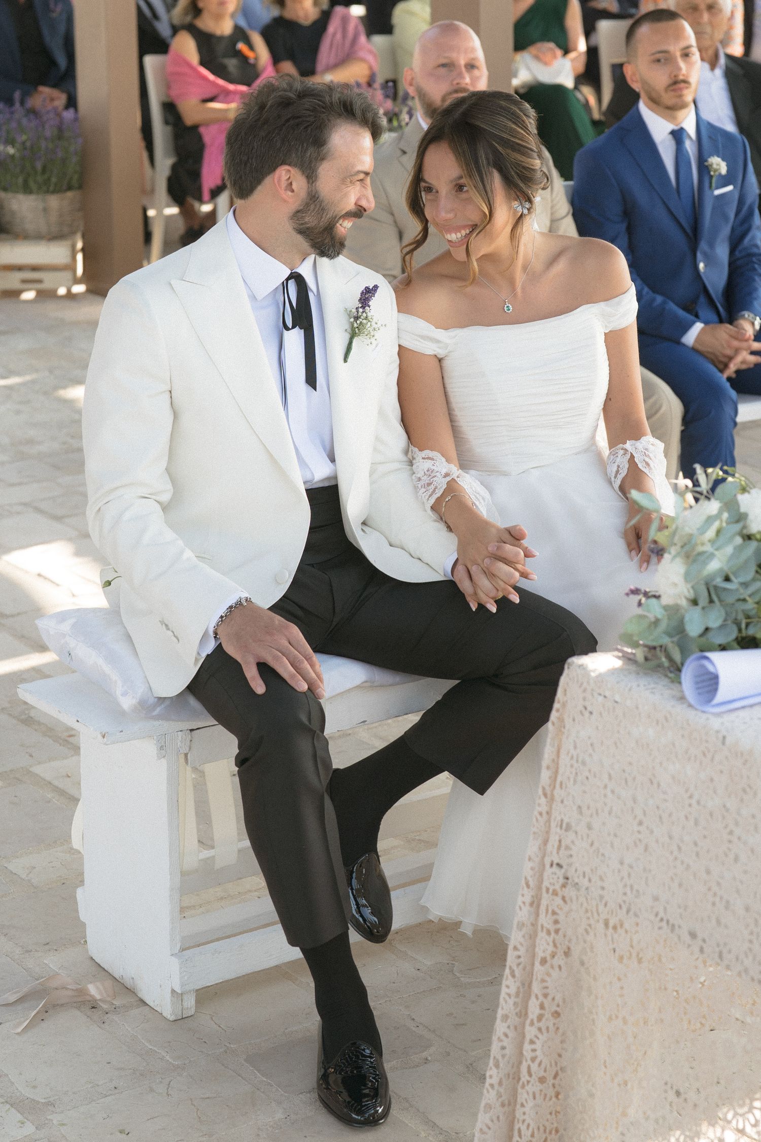 Newlyweds share a romantic moment while seated on a white bench at an outdoor ceremony.