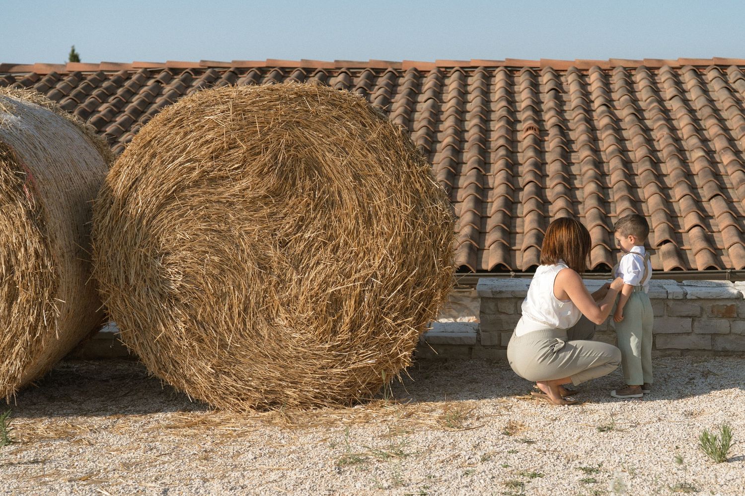 Person sitting beside large round hay bales against a textured roof backdrop in rural setting.