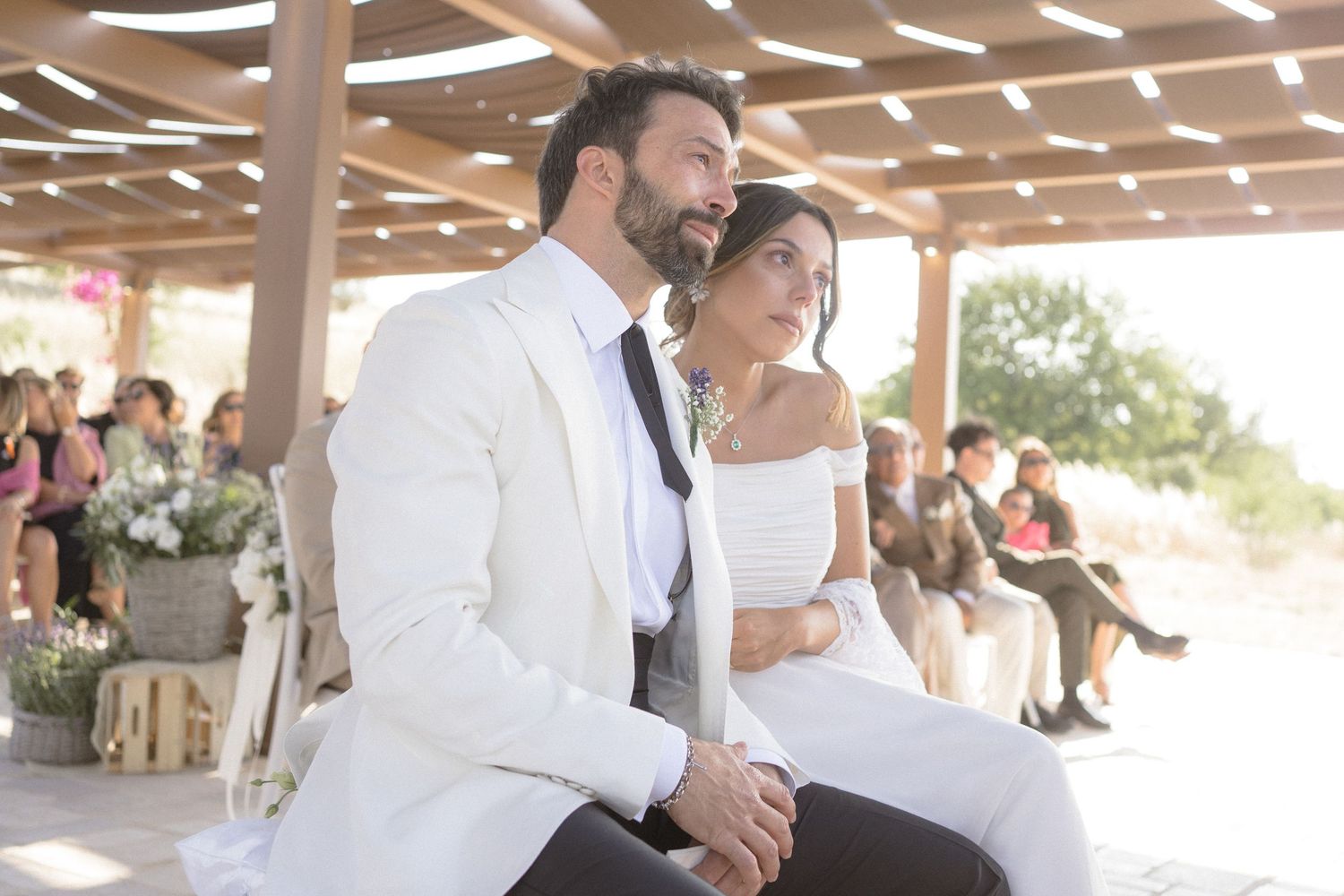 A couple seated together during their outdoor wedding ceremony under a wooden pergola.