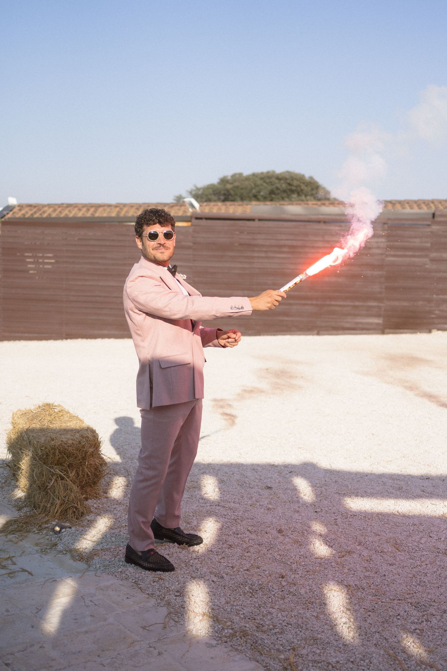 A person in a pink coat holding flares on a rooftop against a bright blue sky.