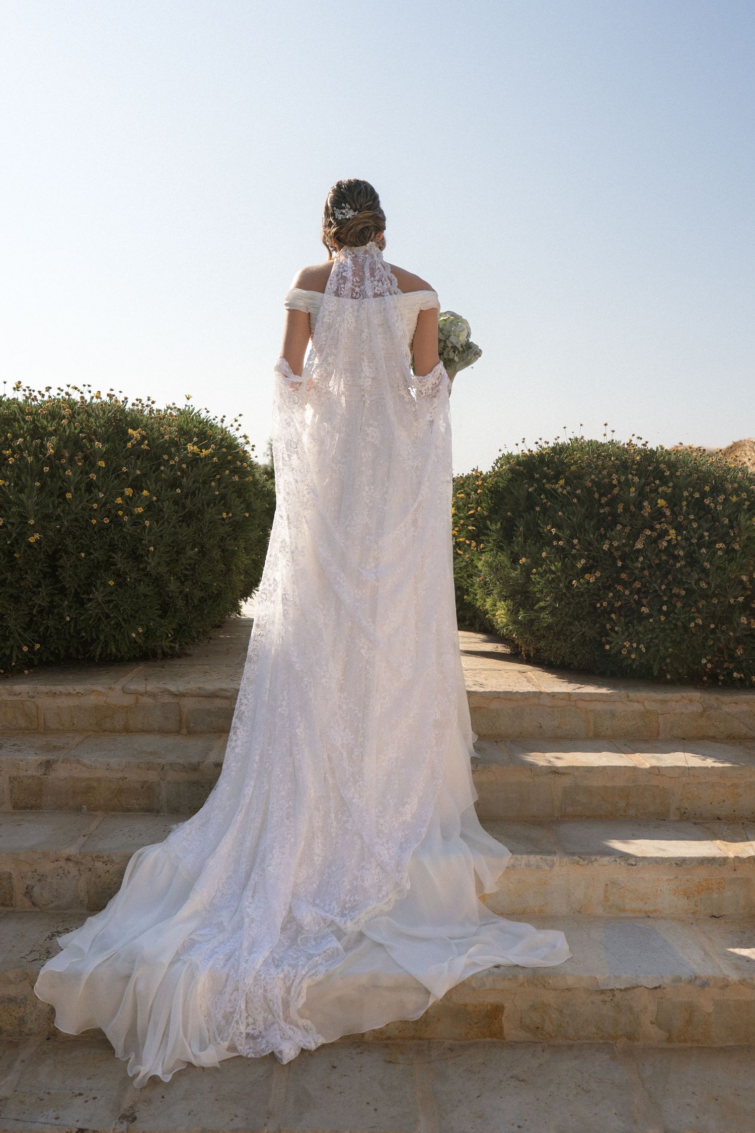 Back view of a bride in flowing lace wedding dress standing on stone steps with garden backdrop.