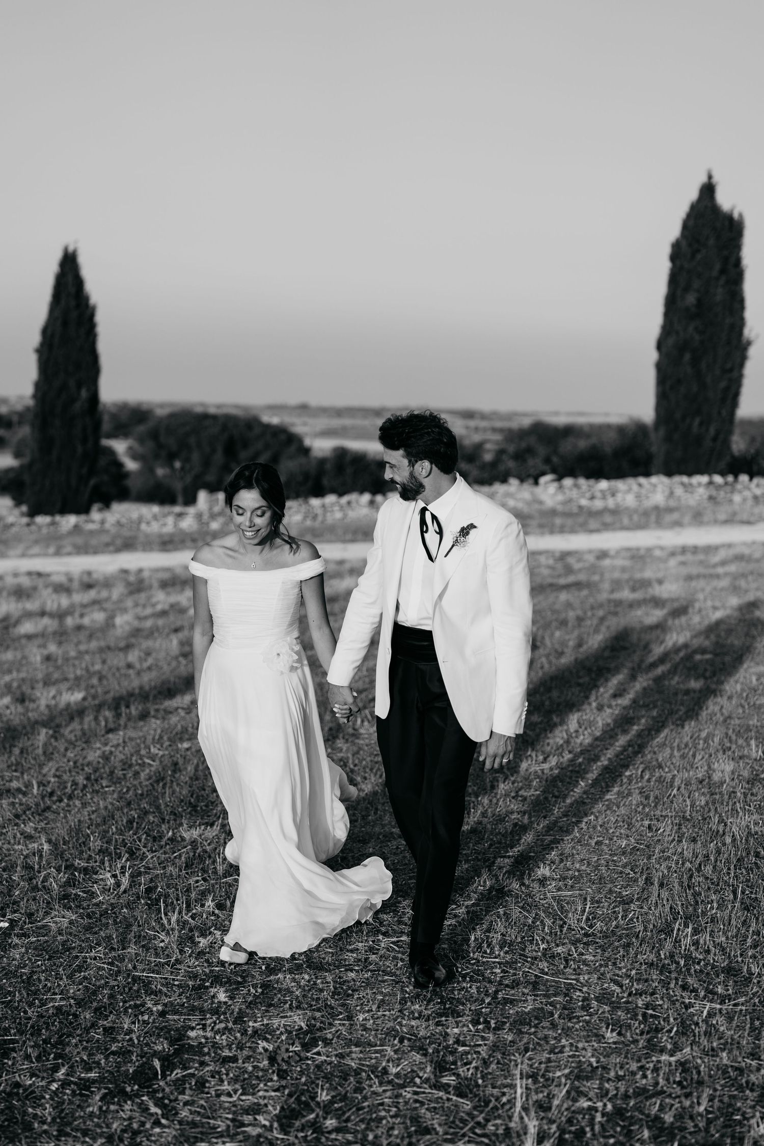 Elegant couple in wedding attire walking through a scenic Tuscan landscape with cypress trees.
