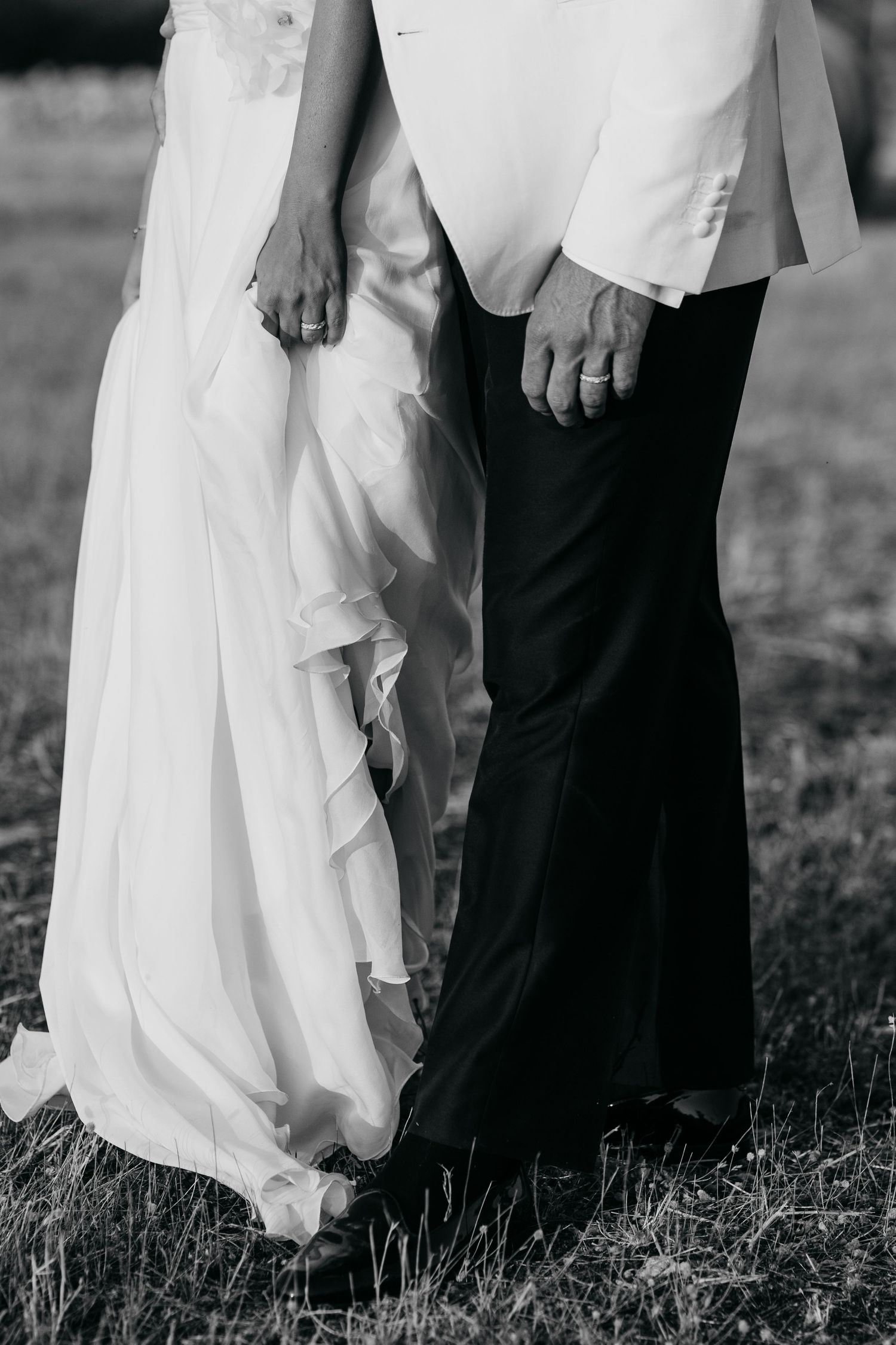 Close-up black and white photo showing feet at a wedding with a flowing white dress and dark formal shoes on grass.