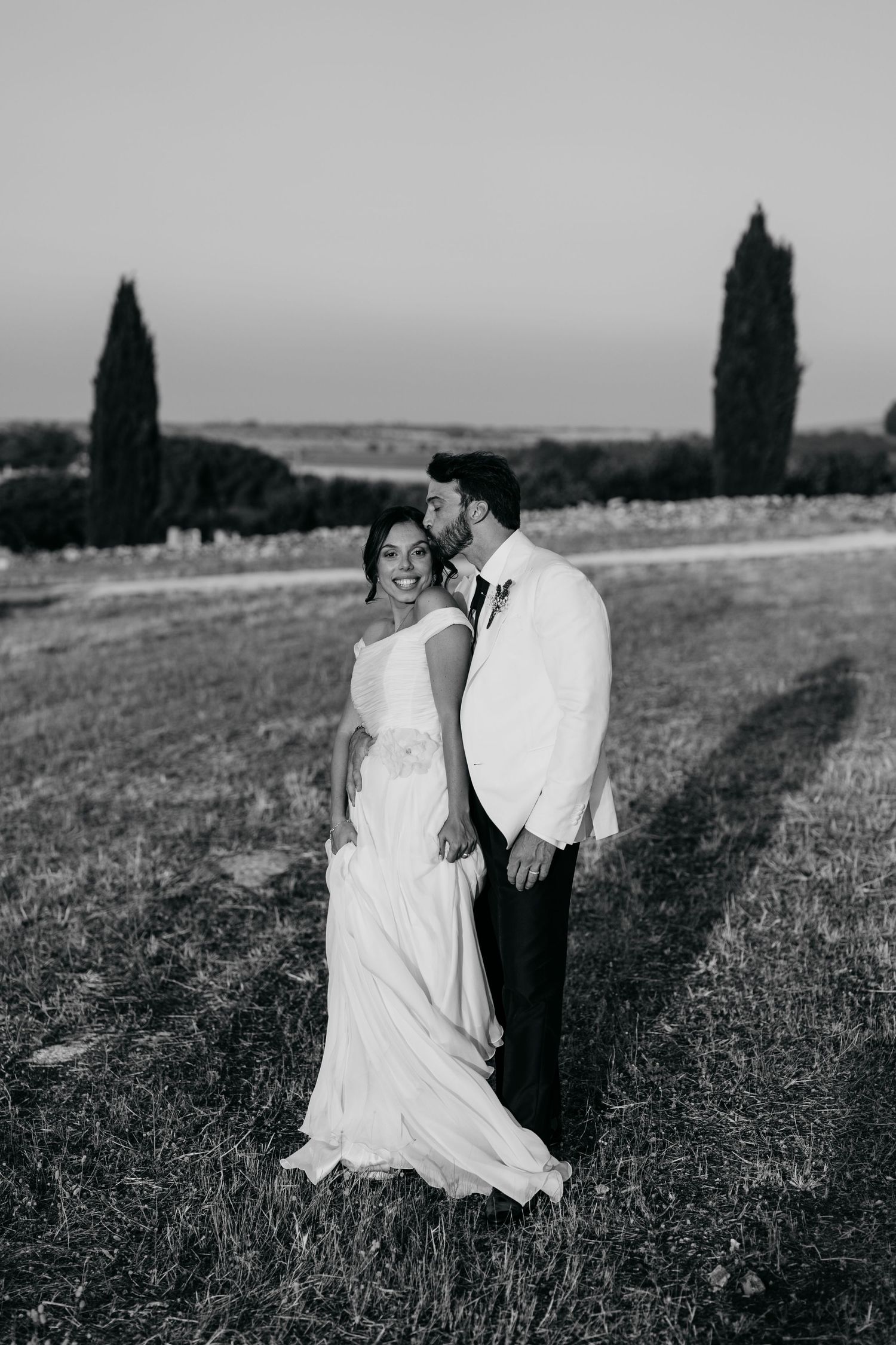 A romantic black and white photo sequence of a couple embracing in a rustic countryside setting with cypress trees.