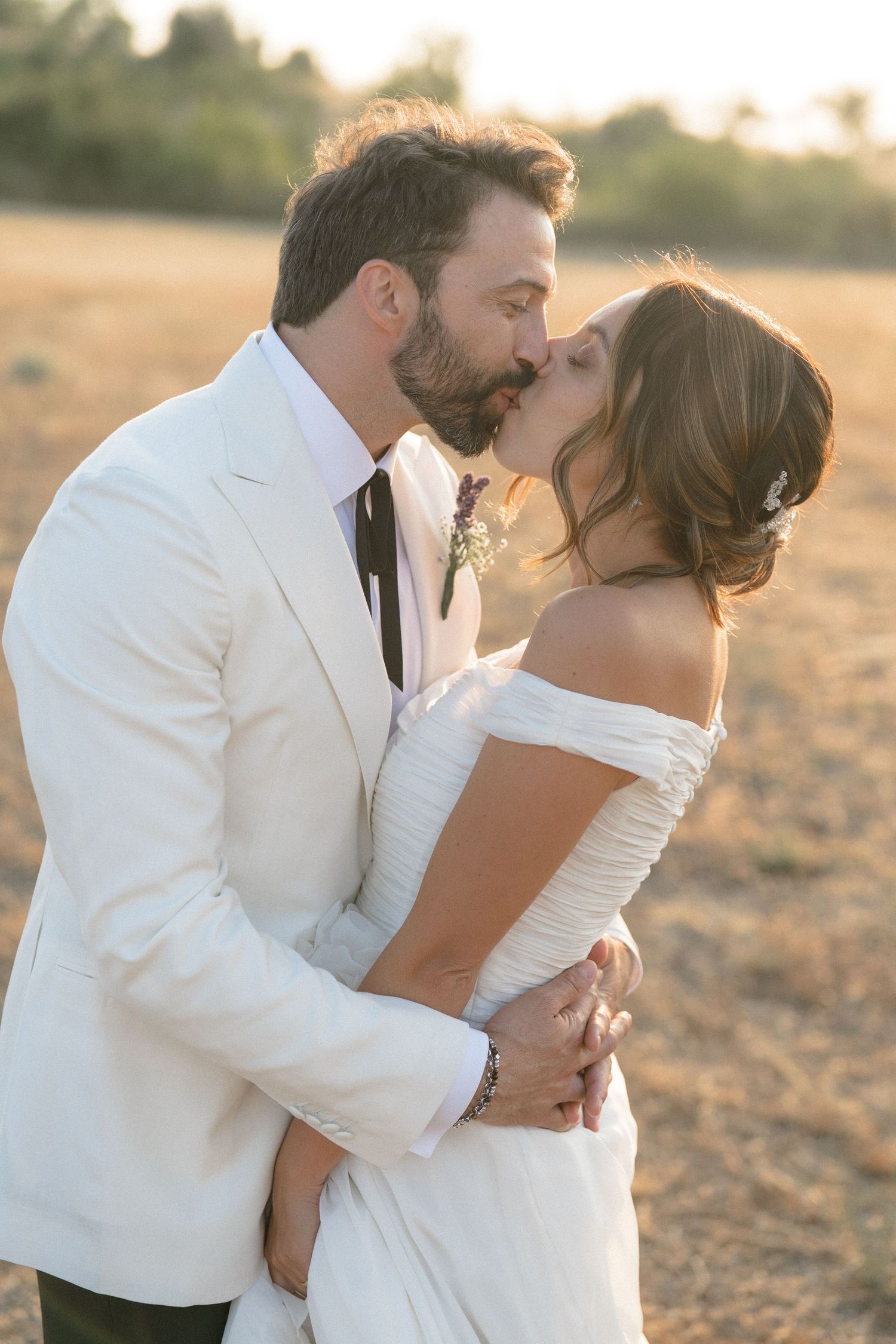 Romantic newlywed couple embraces in golden sunlight wearing elegant white wedding attire.