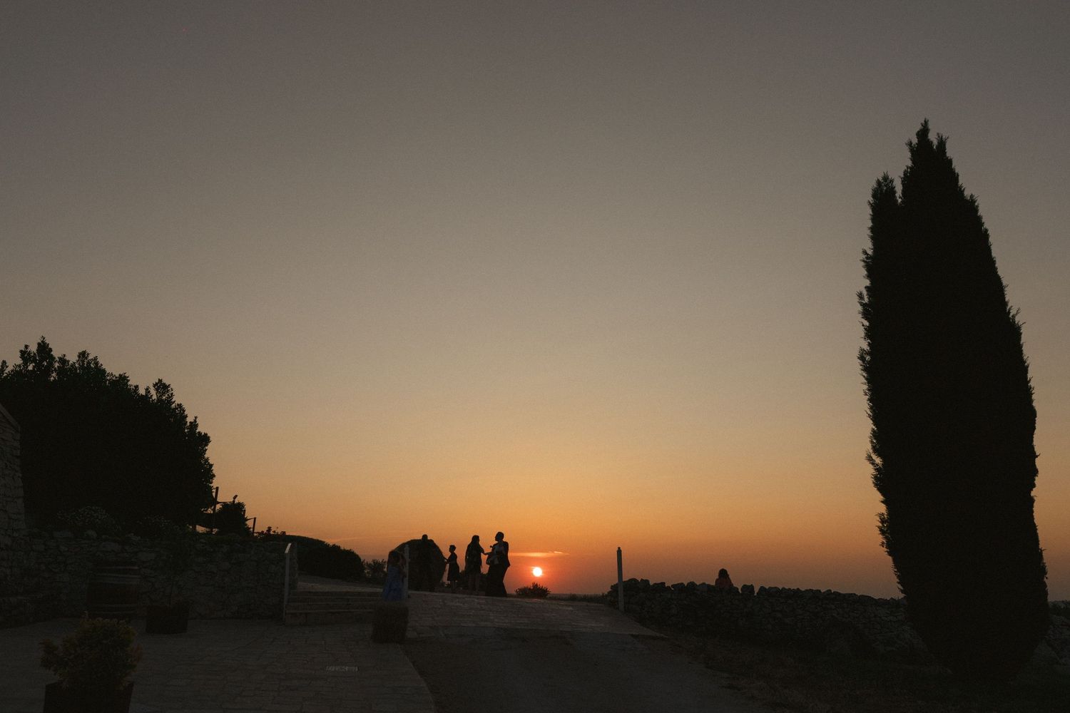 Silhouettes of trees and people against a sunset sky with warm orange and purple hues.