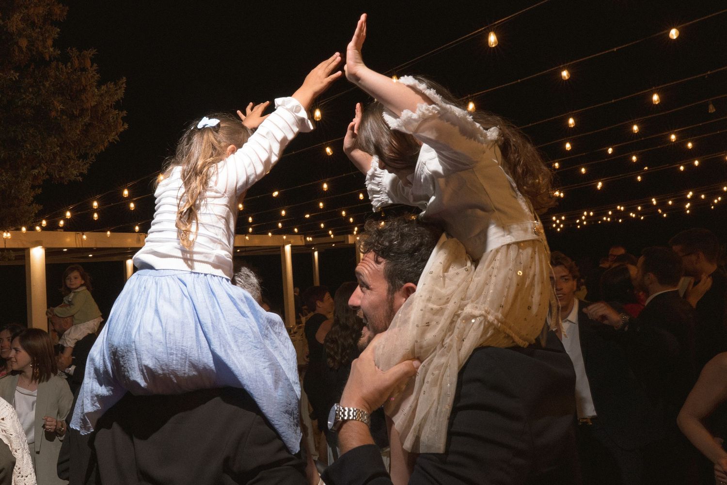 Wedding guests dancing and celebrating beneath string lights at an outdoor reception.