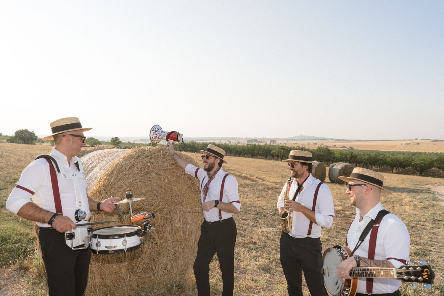 Group of people in wedding attire gather around hay bales in a sunny rural field.