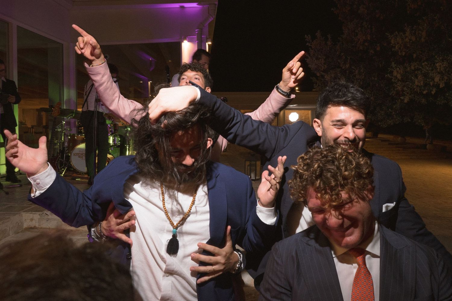 Wedding guests dancing and celebrating enthusiastically on the dance floor at a nighttime reception party with purple lighting.
