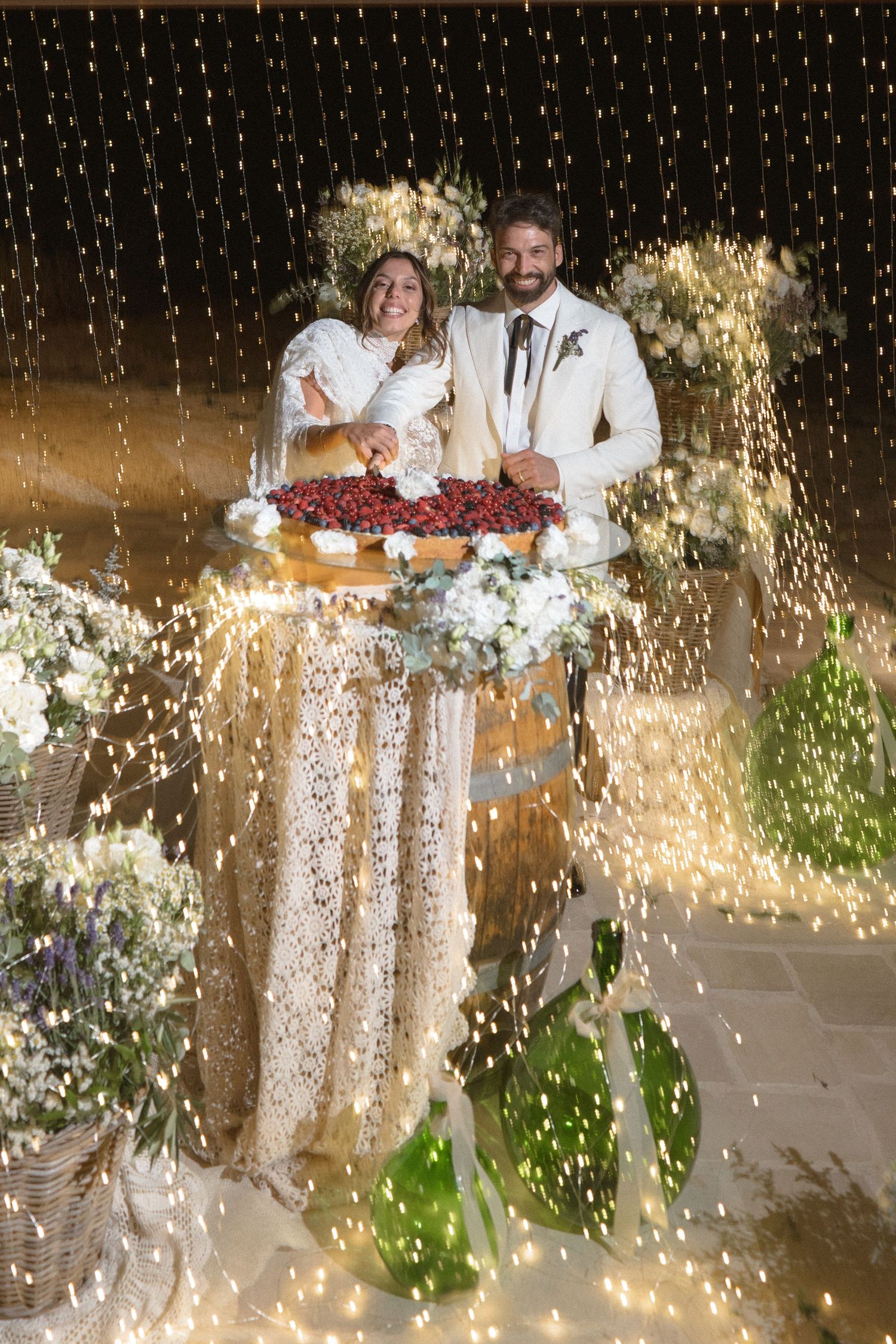 Couple cuts wedding cake surrounded by sparkling lights and floral decorations at an outdoor evening reception.