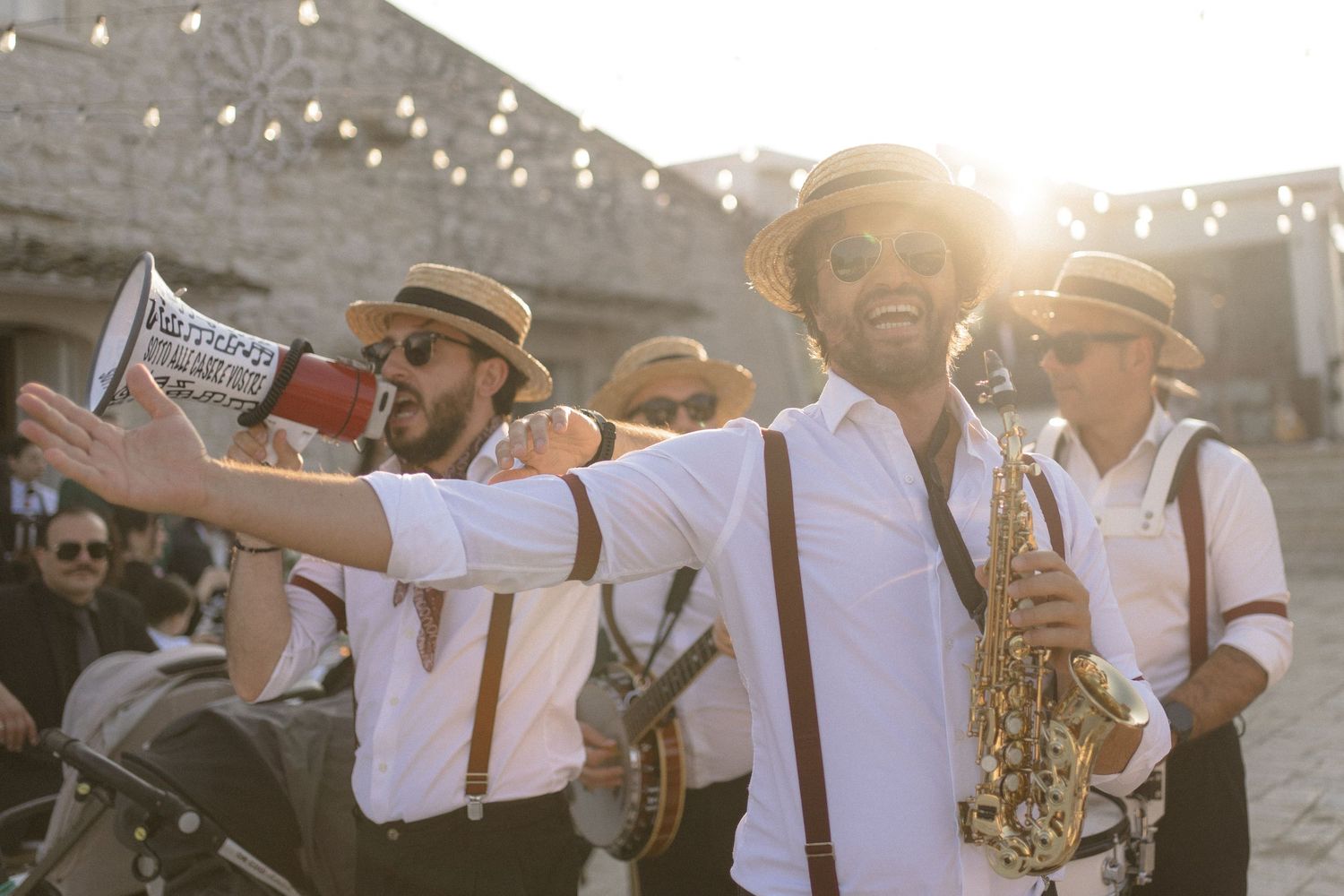 Band members in white shirts and suspenders playing instruments outdoors under string lights at sunset.