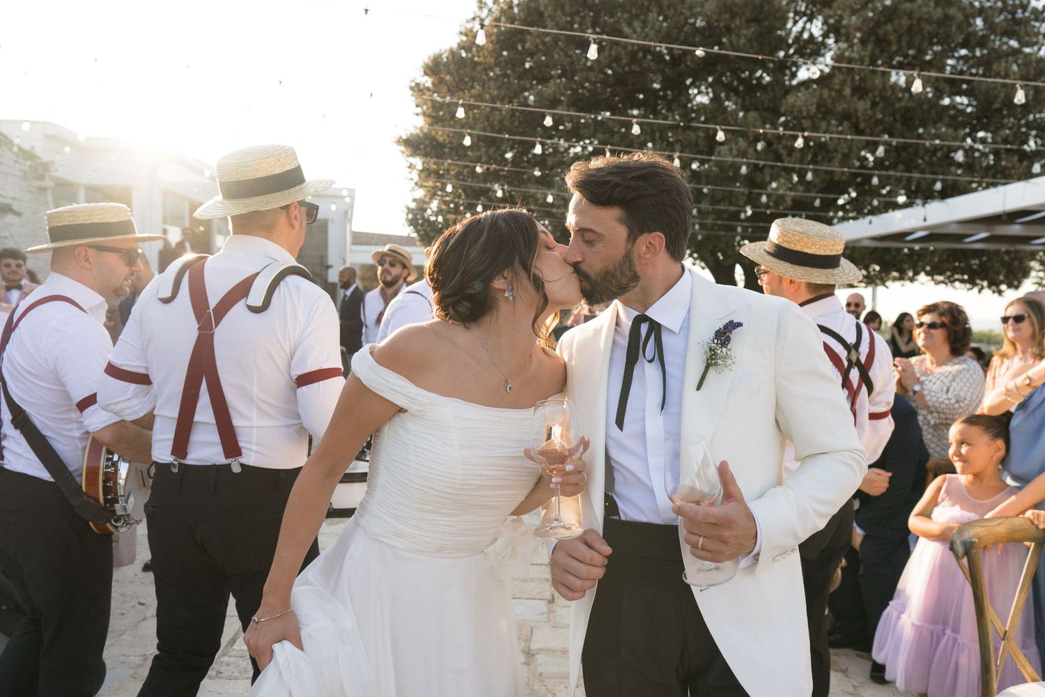 Couple dancing at outdoor wedding reception with guests in suspenders and straw hats in background at sunset.