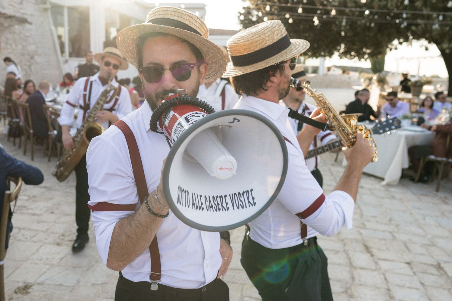 Musicians in vintage attire perform with megaphone and instruments at an outdoor wedding.
