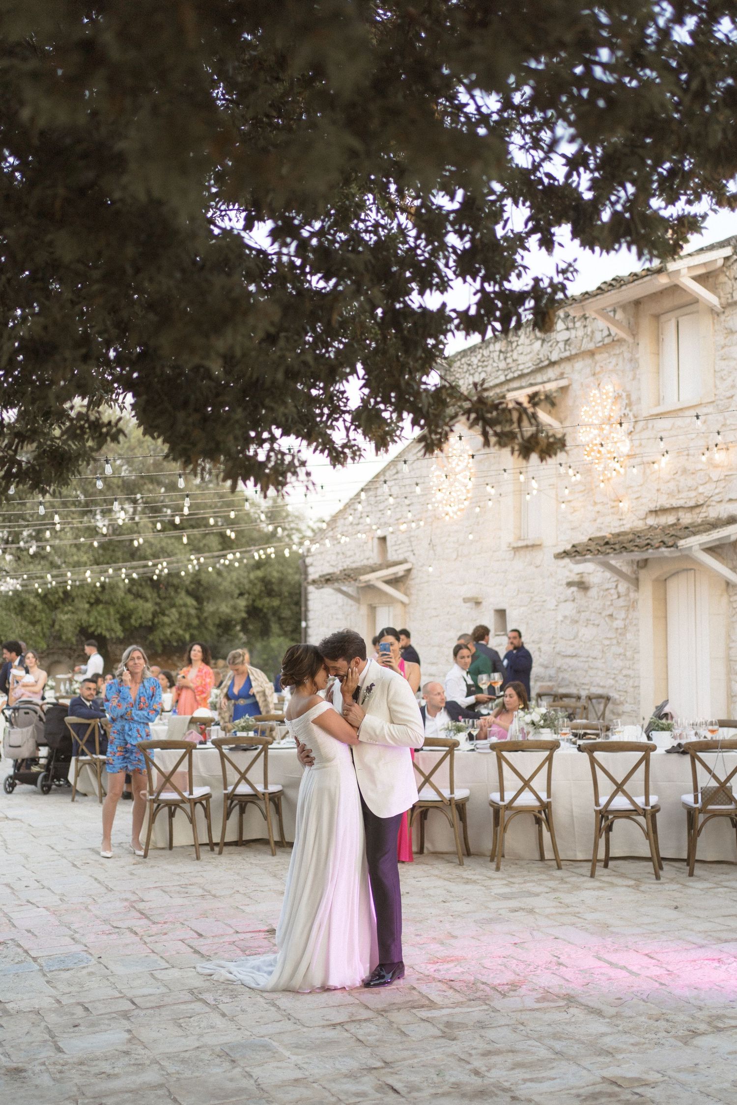 A couple dances during a romantic outdoor wedding reception with string lights and rustic stone walls.