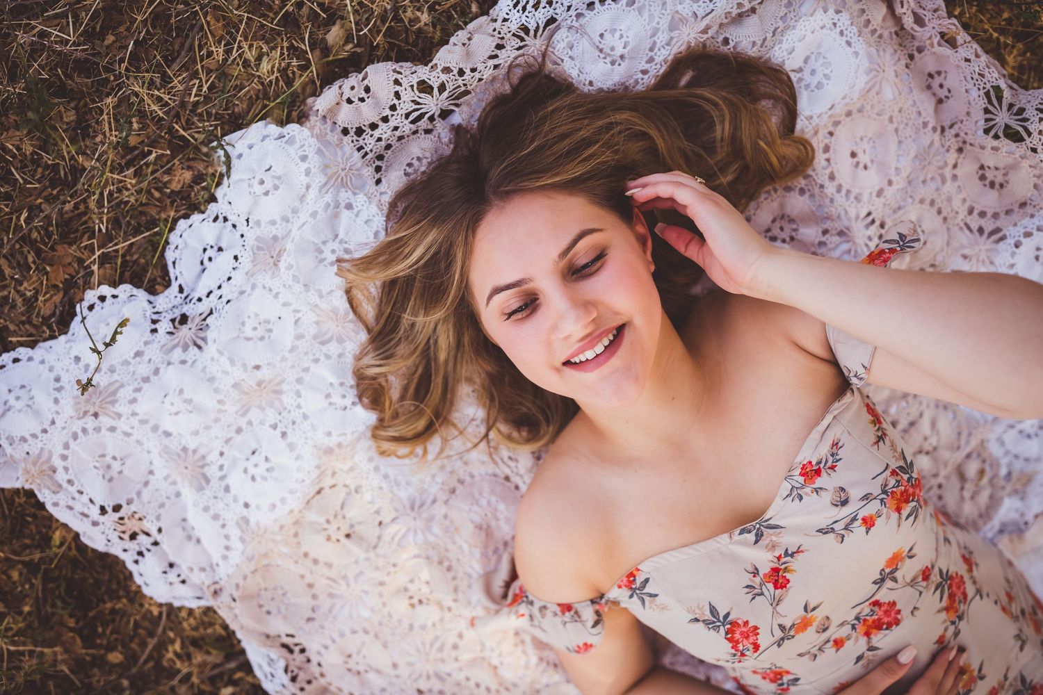 A joyful young woman in a floral dress lies on delicate white lace while resting on natural ground.
