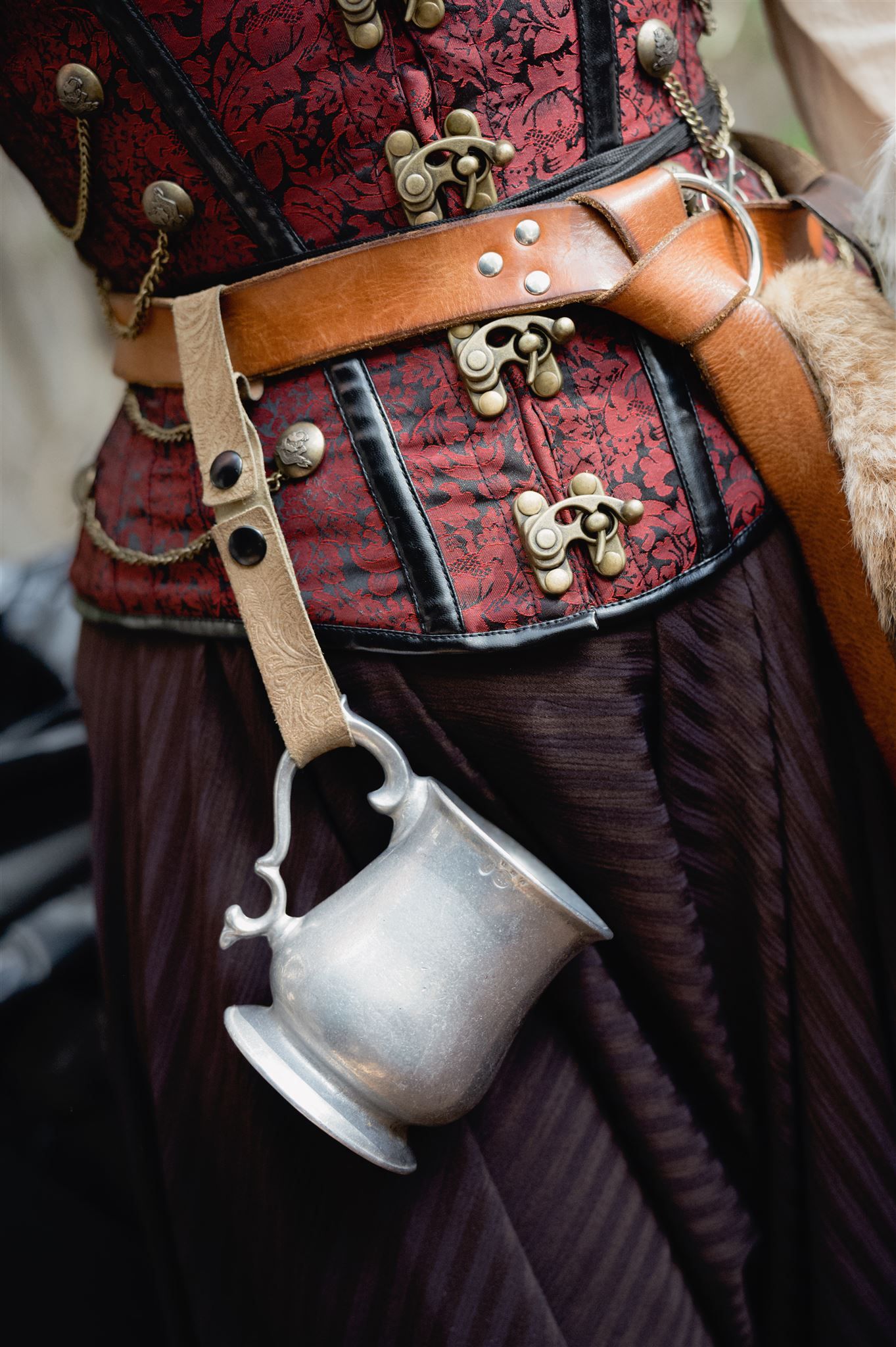 Medieval style leather belt and corset with ornate brass clasps and hanging pewter tankard against red and black fabric.