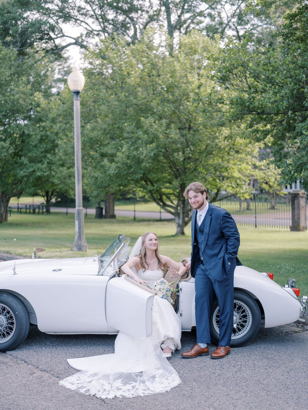 Classic white vintage MG convertible sports car parked under trees with couple in wedding attire standing beside it.