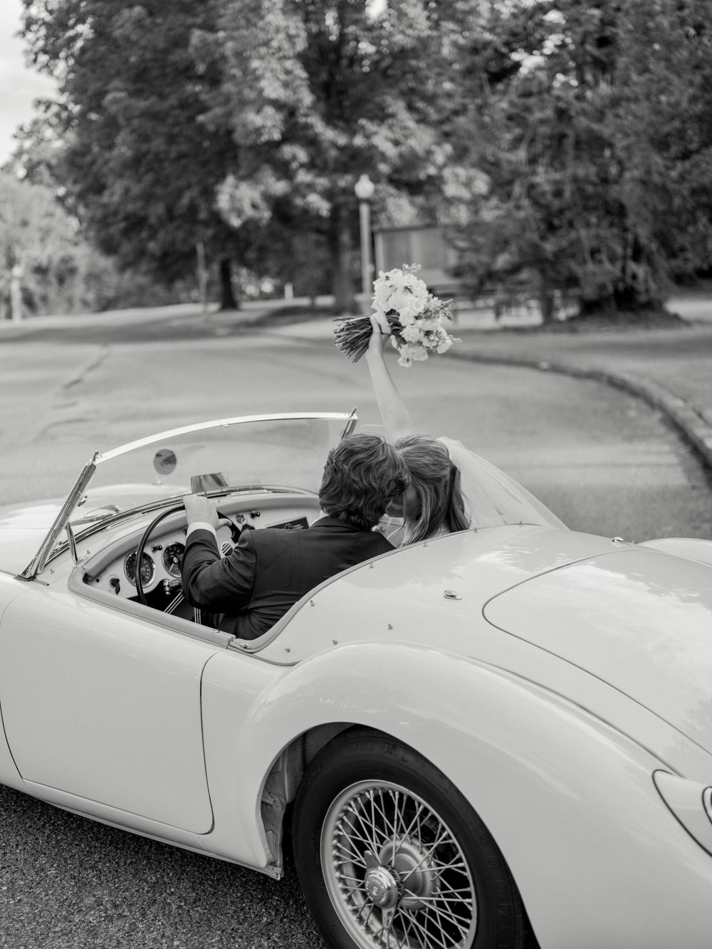 Black and white photo of a classic convertible sports car with wire wheels parked on a tree-lined road.