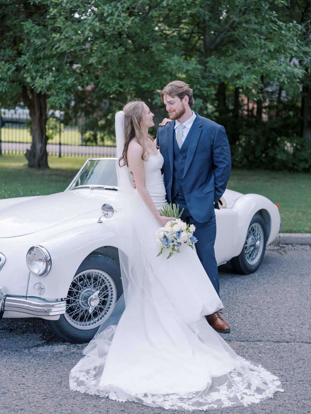 A couple poses with a classic white vintage car during their wedding, featuring a flowing lace gown and navy suit.