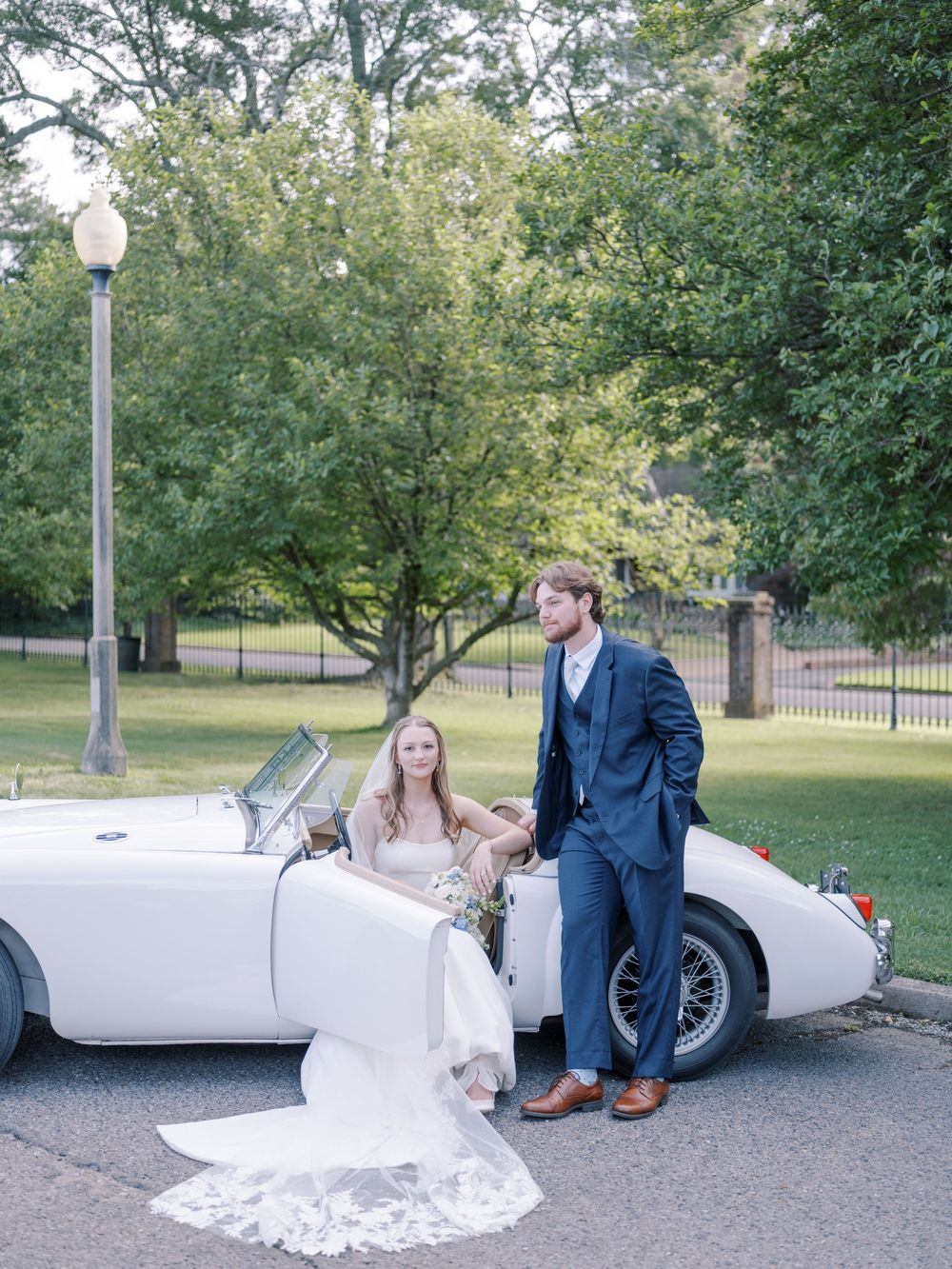 A couple poses with their classic white vintage convertible car in a park setting with elegant wedding attire.