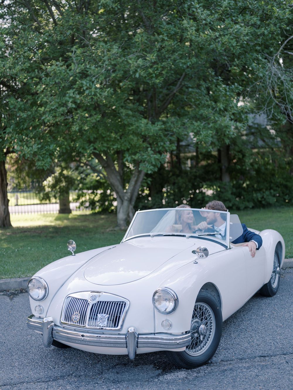 Classic white MG convertible sports car parked on a driveway under green trees on a sunny day.