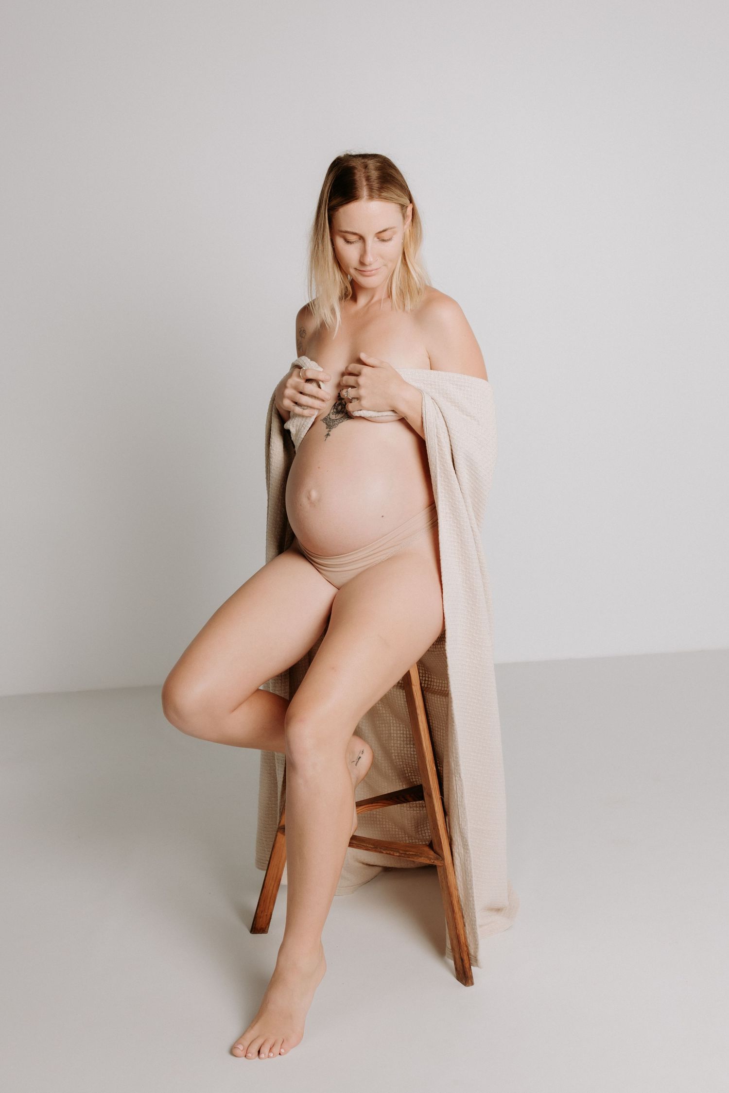 Minimalist maternity photo of an expectant mother in soft beige tones sitting on wooden stool against neutral background.