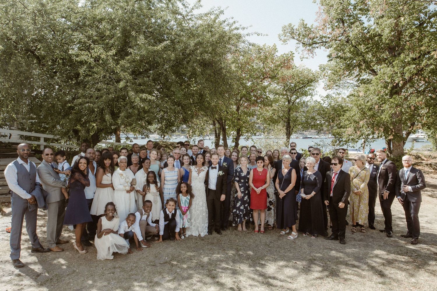 Large wedding party poses together under shady trees for a group photo.