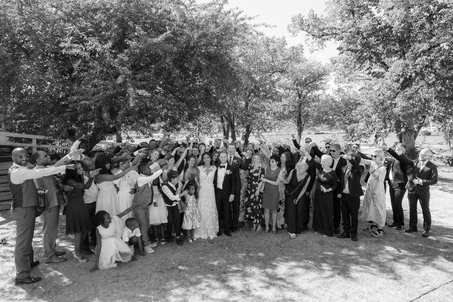 Black and white photo of large wedding party gathered outdoors under shaded trees.