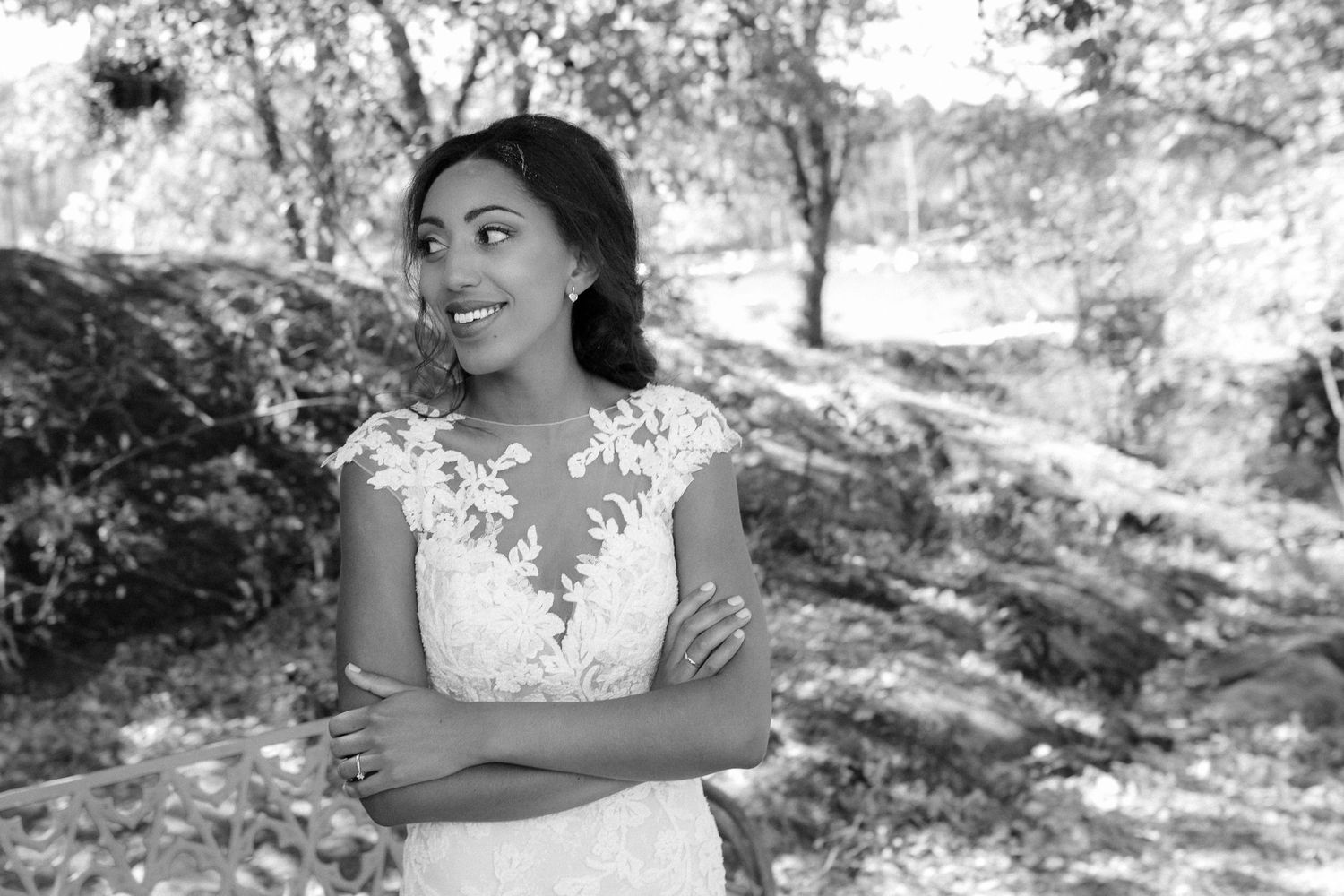 A bride in a lace detailed dress stands with crossed arms in a black and white outdoor portrait.