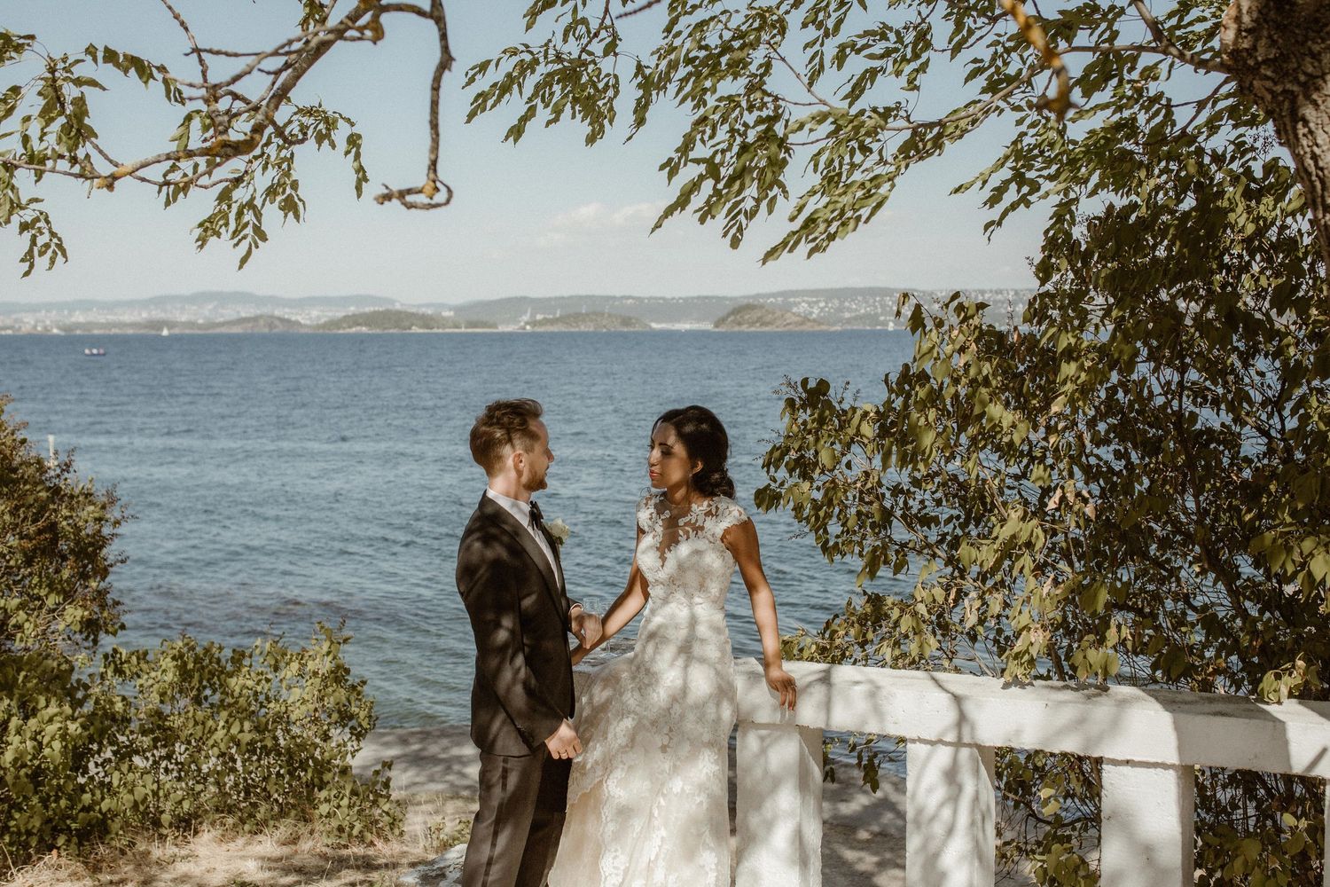 A couple in wedding attire stands together on a scenic ocean overlook with a white railing and overhanging tree branches.