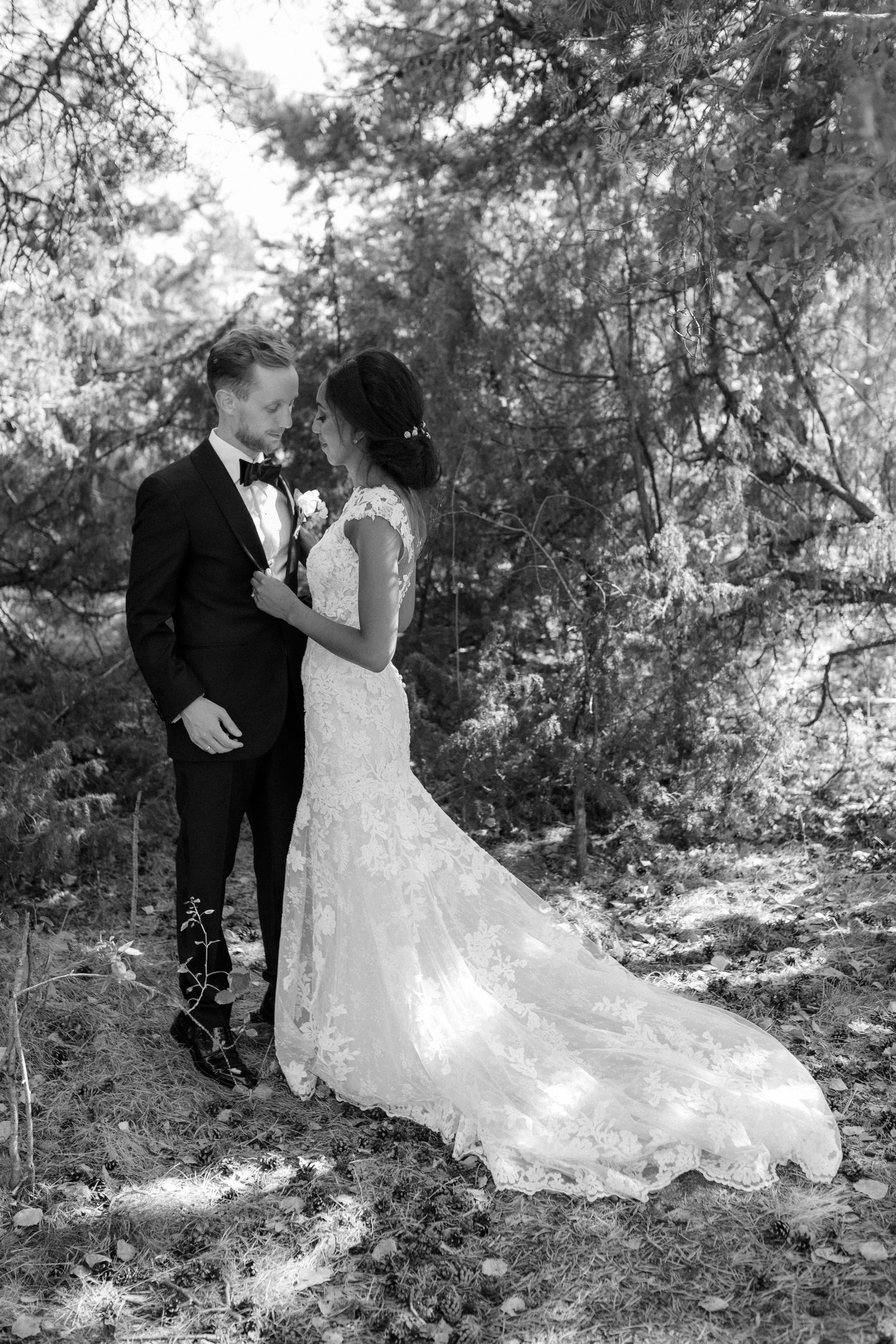 A romantic black and white wedding photo of a couple in formal attire embracing in a wooded outdoor setting.