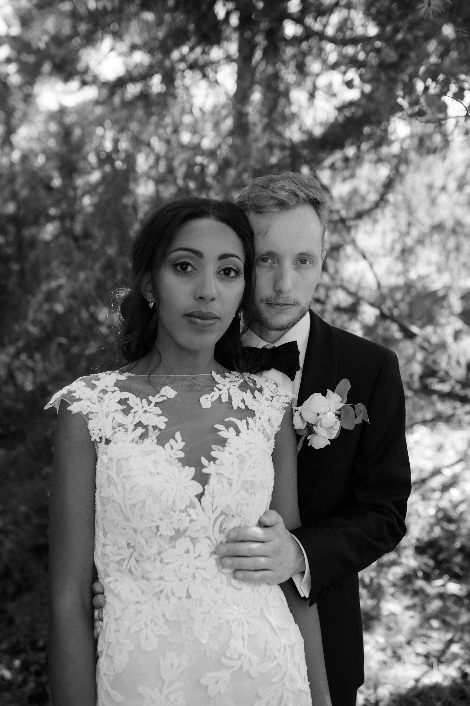A black and white portrait of a bride in lace gown and groom in tuxedo standing together in a forest setting.