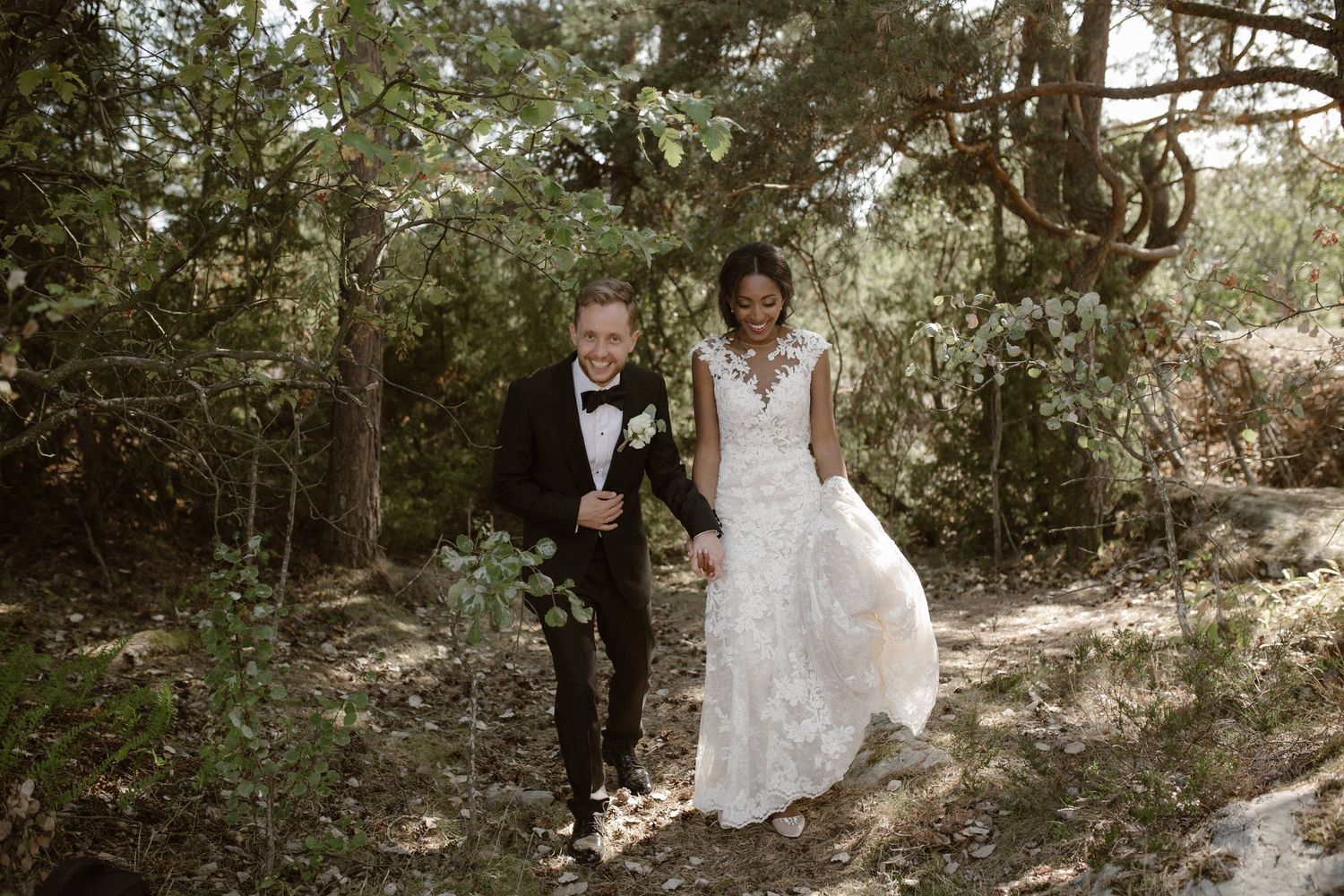 Couple in wedding attire walks hand in hand through a wooded outdoor setting.