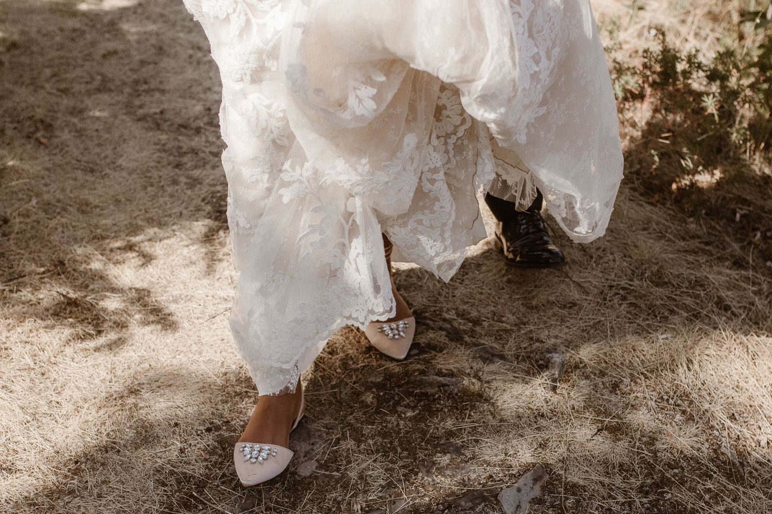Close-up view of white lace wedding dress hem and bridal shoes on rustic ground in sepia tones.