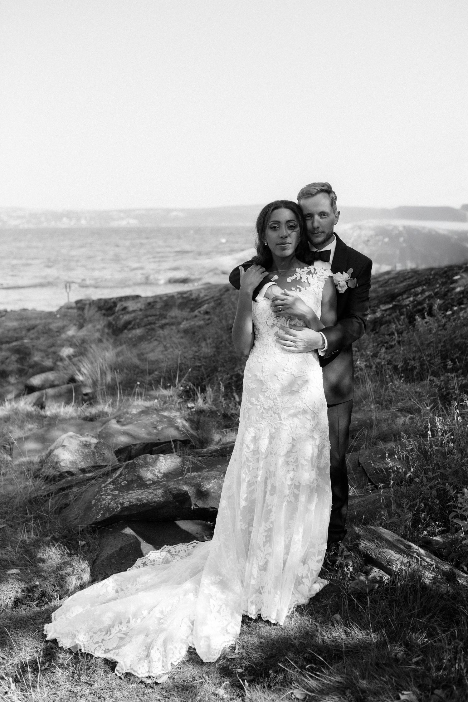 Black and white portrait of a couple in wedding attire standing on a coastal hillside.