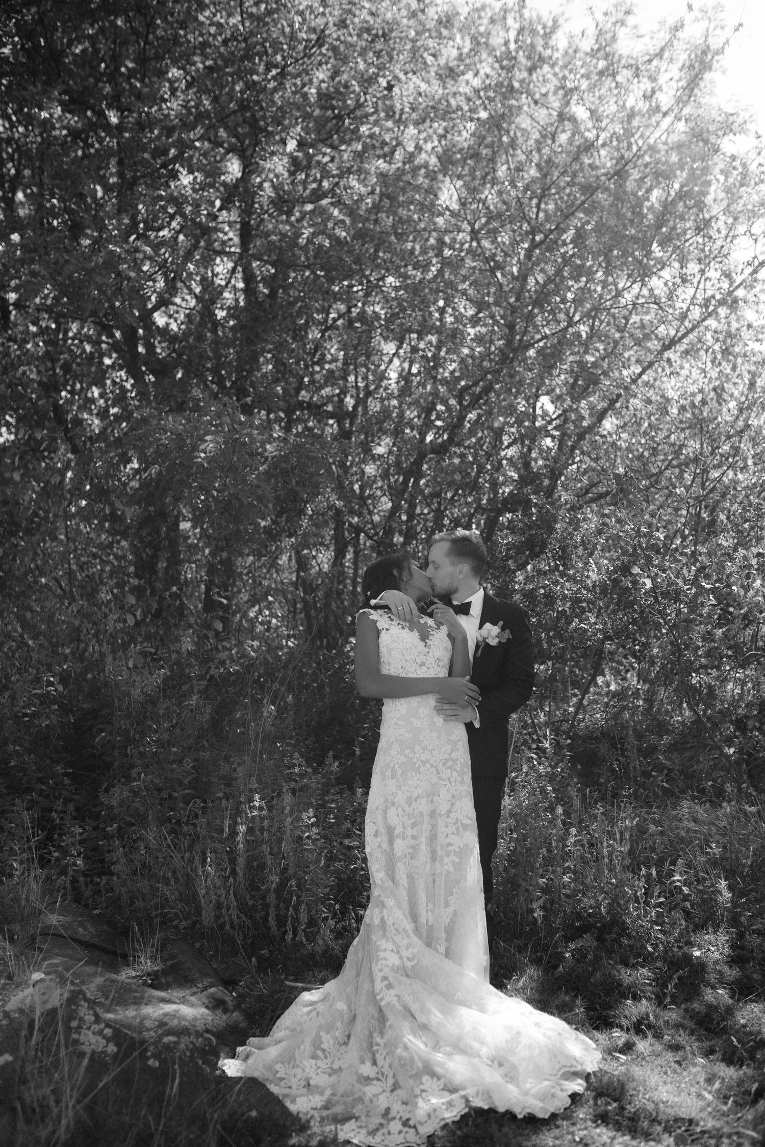 A romantic black and white wedding photo showing a couple embracing among trees in an outdoor setting.