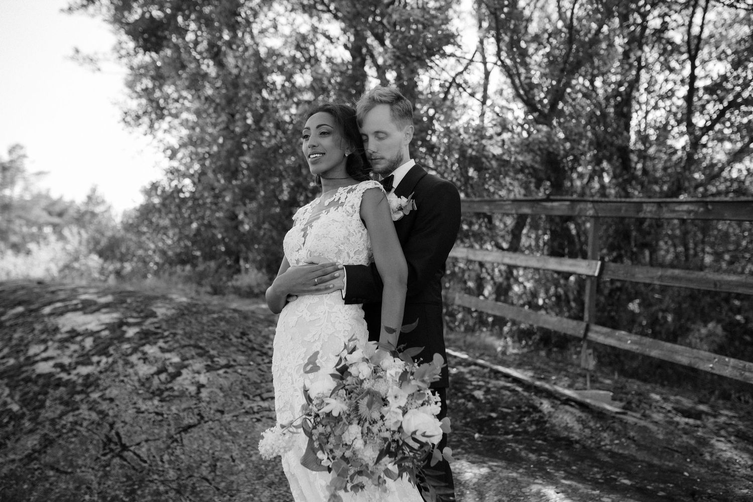 A black and white wedding photo of a couple standing together on a rustic wooden bridge surrounded by trees.