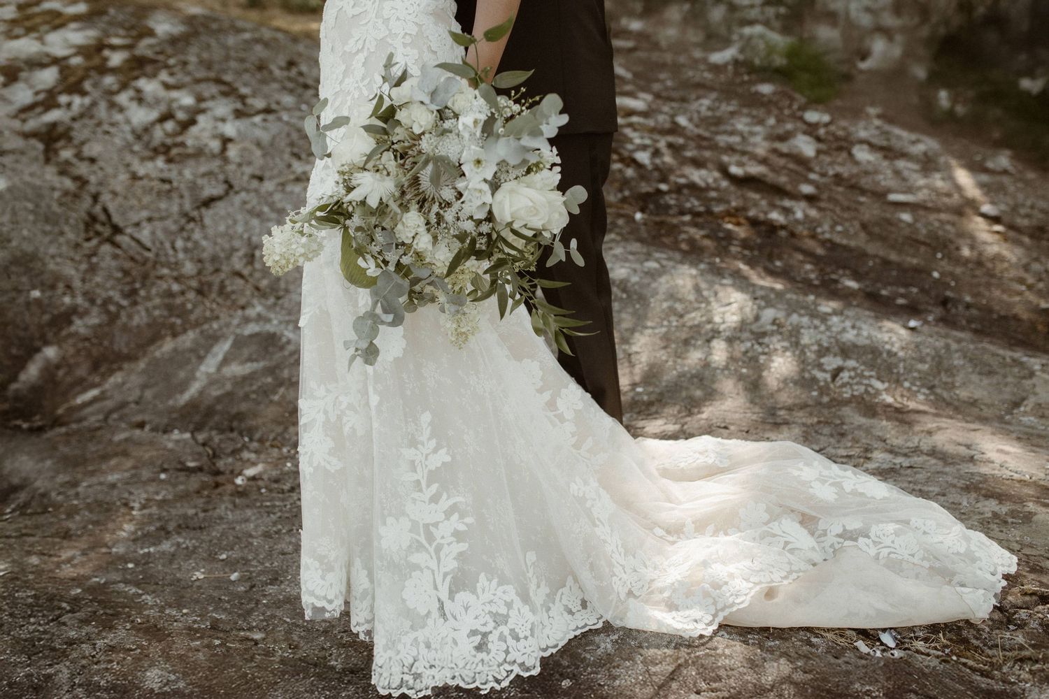 Close up of an elegant lace wedding dress train and white bridal bouquet against a rocky backdrop.